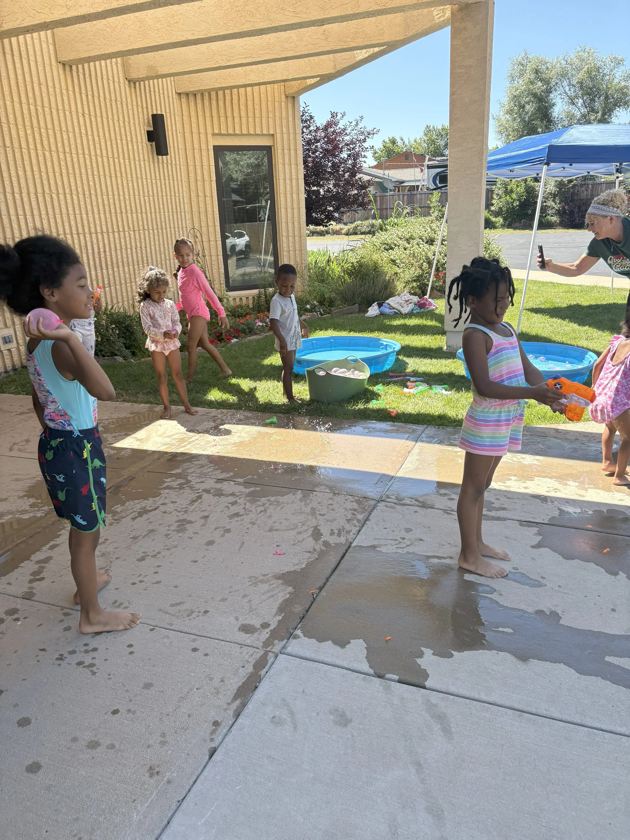 Children playing with water guns and splashing water on a patio during a sunny day, with a person taking photos