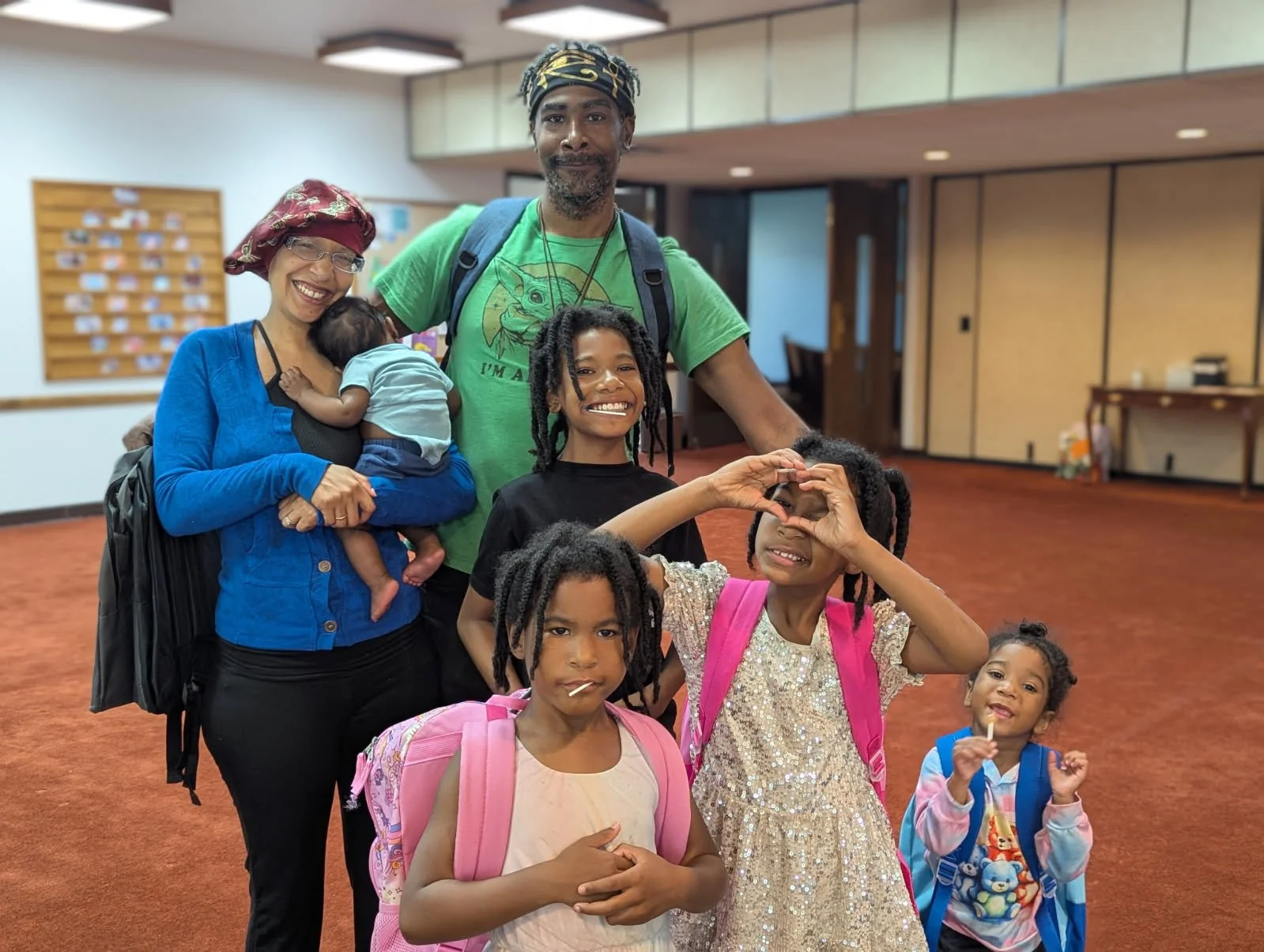 Group of five children and two adults, smiling inside a building with a wooden wall and red carpet, some children have backpacks, one child makes a heart shape with her hands, others hold lollipops.