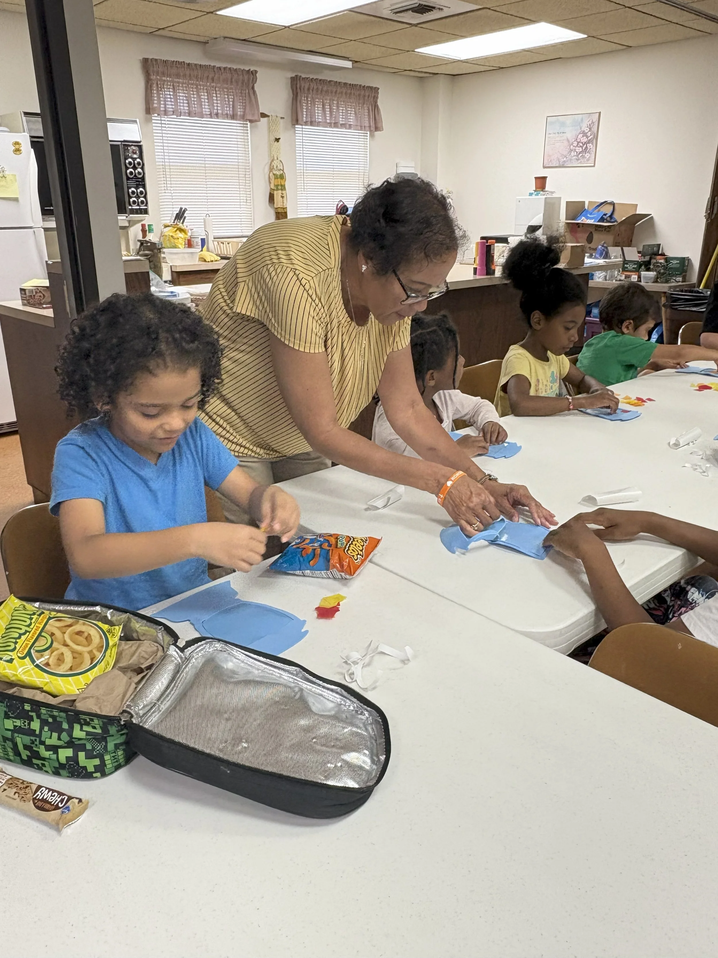 An older woman and several children are sitting at a table, engaging in arts and crafts activities, with snacks on the table.
