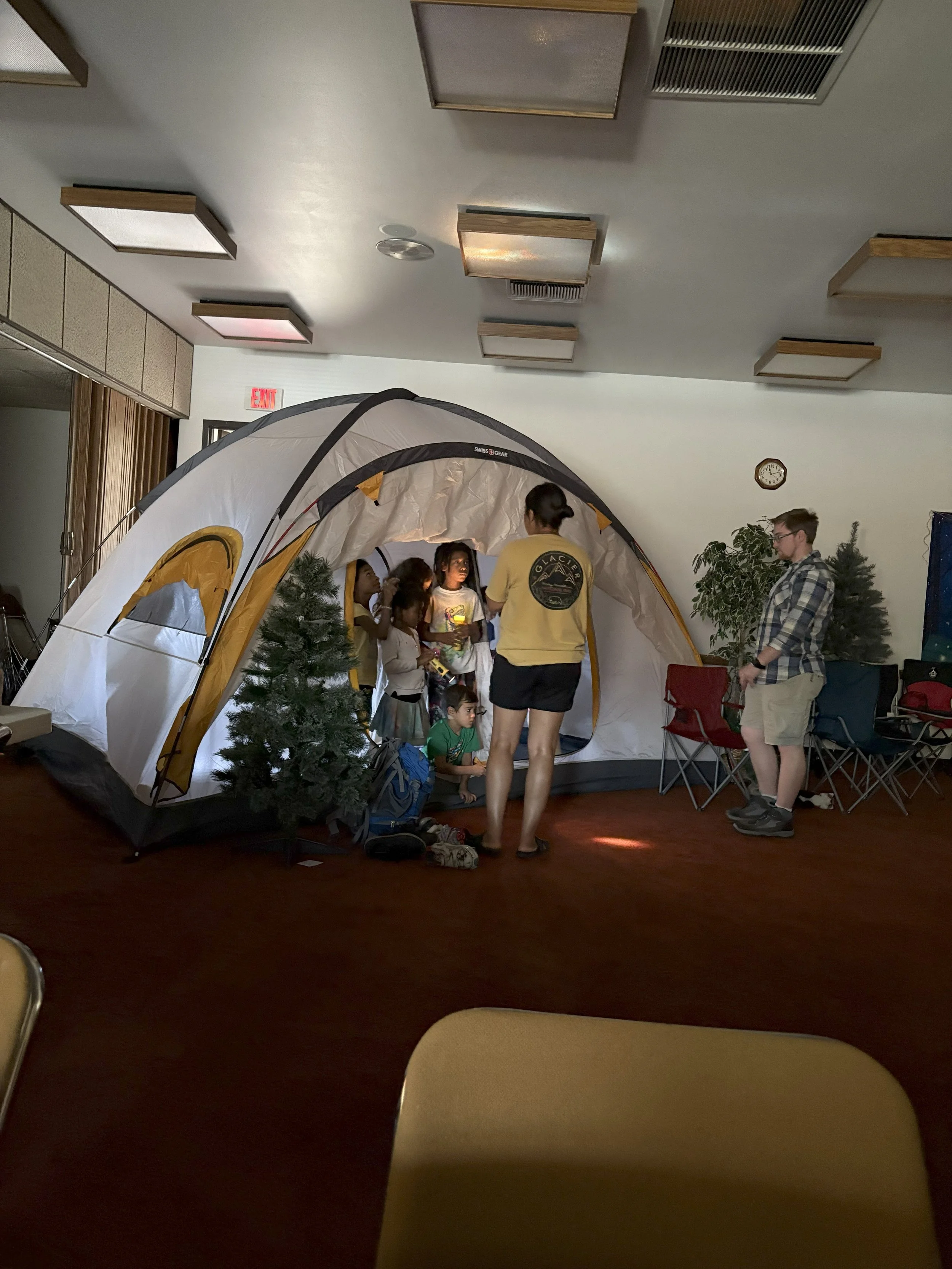 Children and adults gathered inside a large indoor tent, resembling a campsite, with two small artificial trees nearby and chairs around the perimeter.