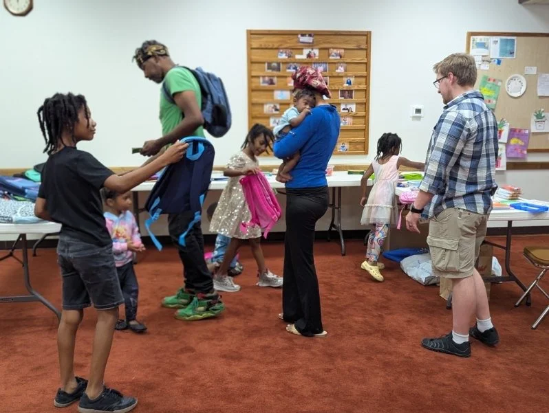 Group of children and adults in a community room, some children are looking at items on tables, while one adult holds a small child and another is looking at a device. The room has a bulletin board, photos, and various supplies on the tables.