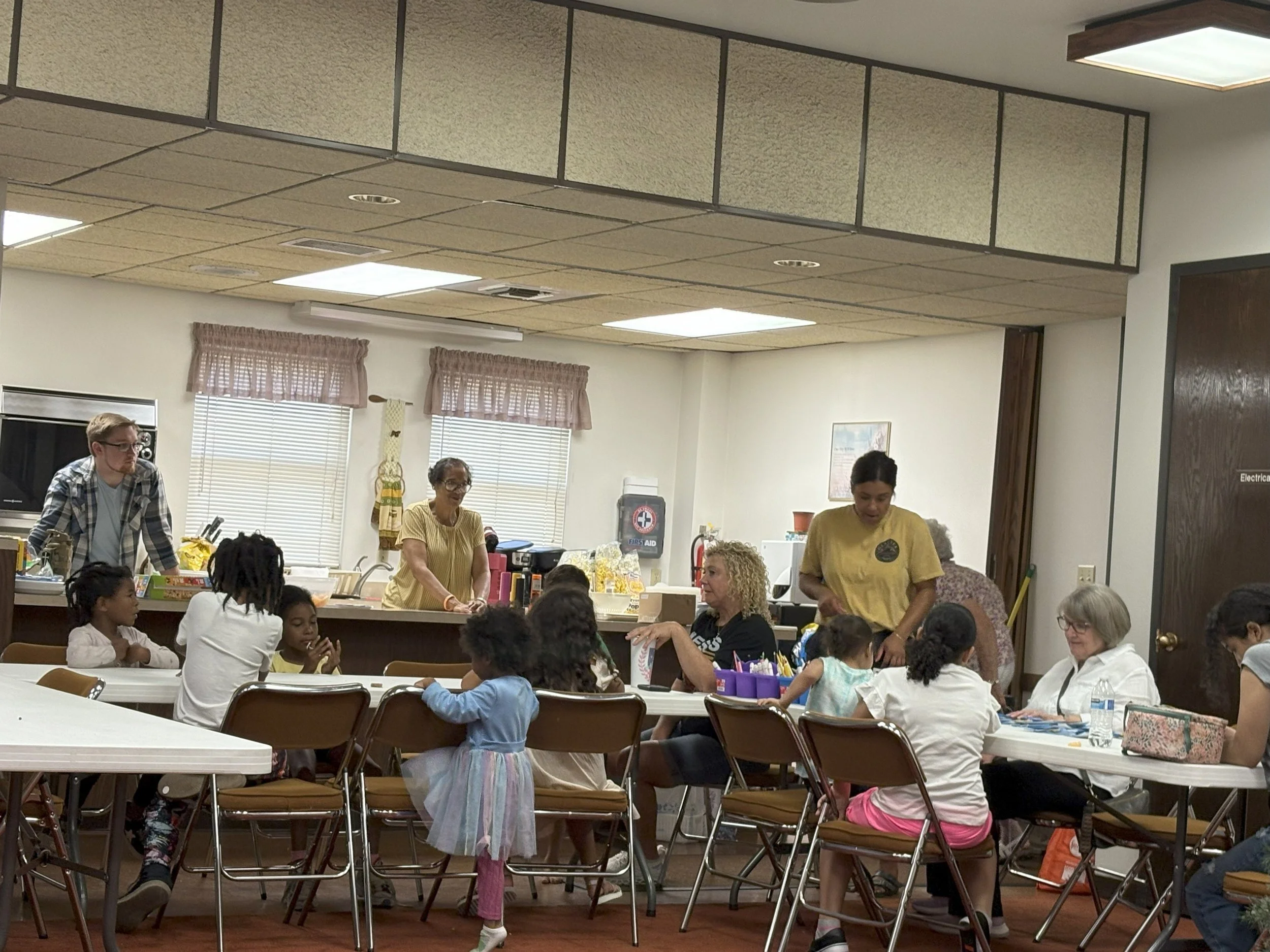 Children and adults in a community room, participating in an indoor activity at tables, with some standing behind the tables and others seated.