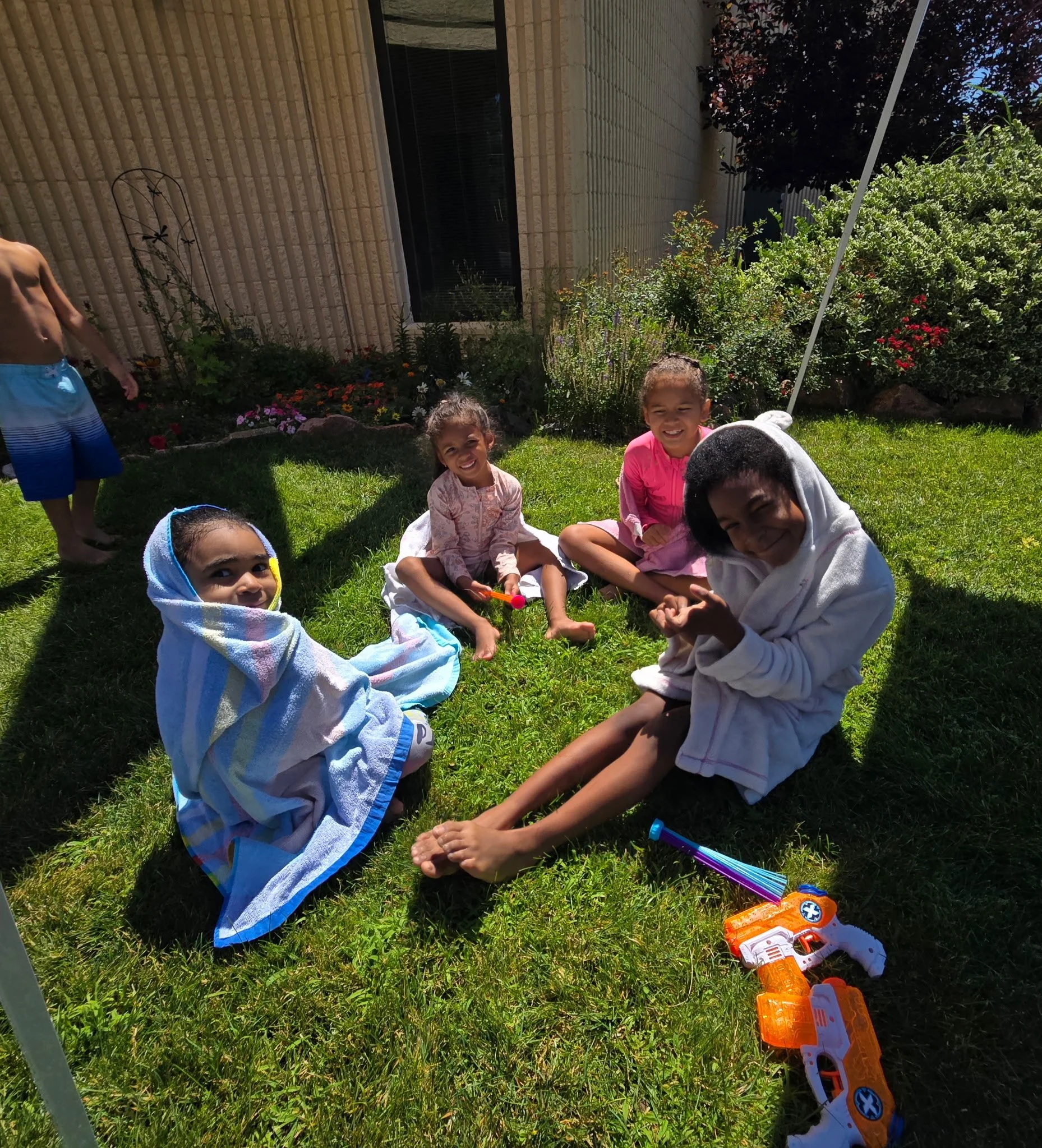 Four children sitting on grass in a backyard, smiling and playing with water toys, with a boy standing in the background and colorful flowers and shrubs nearby.