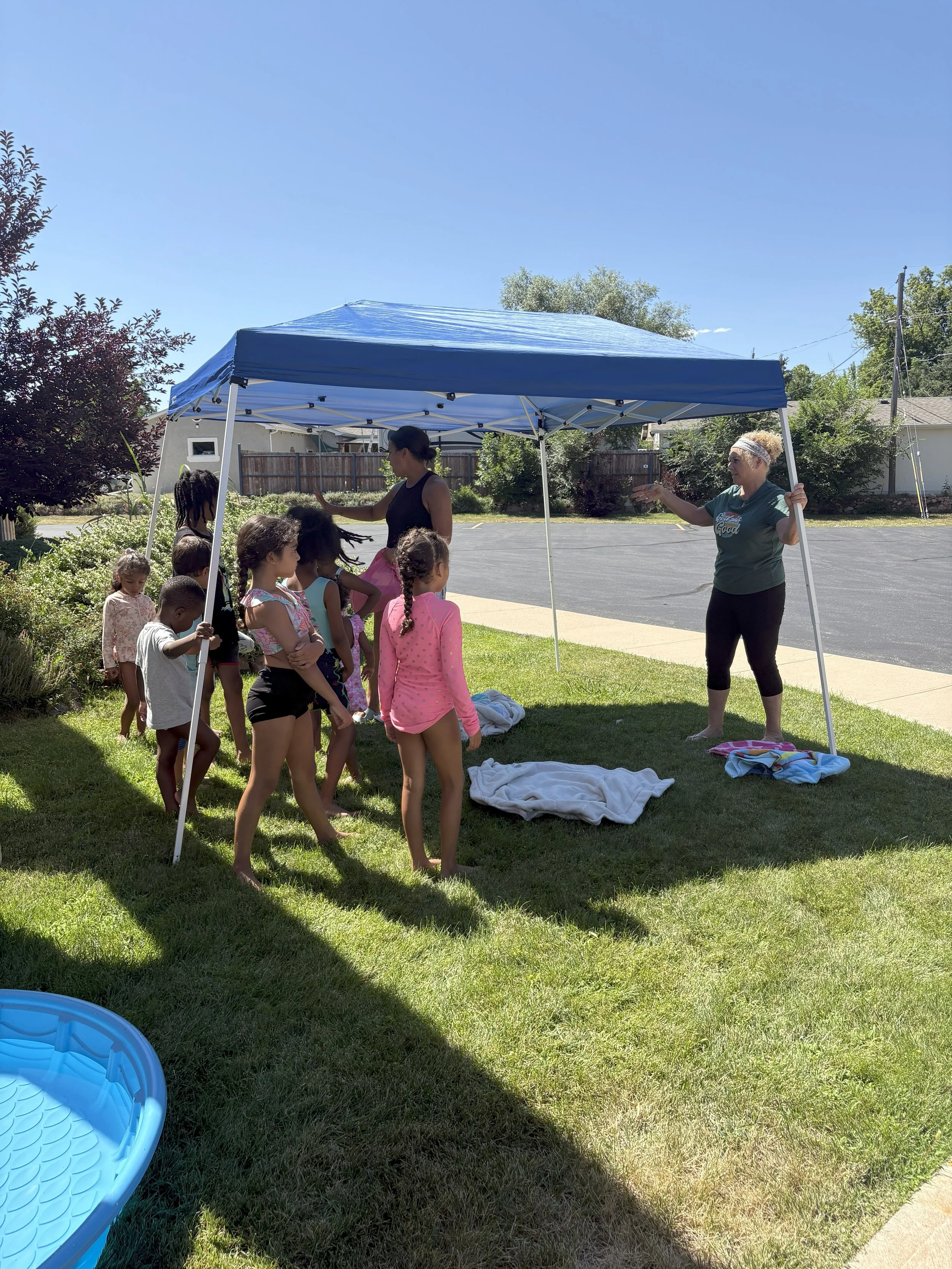 Group of children listening to two women under a blue canopy in a grassy yard on a sunny day.