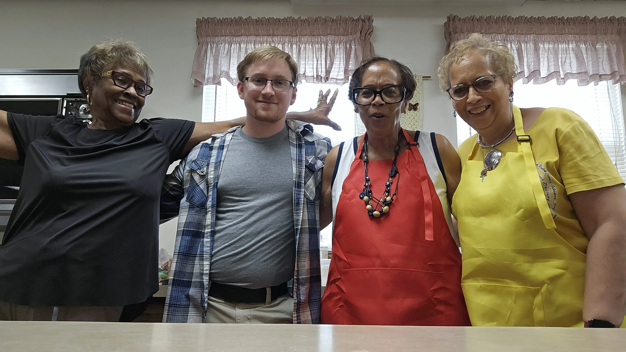 Four mature women and one young man standing together in a kitchen, smiling at the camera. The women are wearing colorful aprons and the man is casually dressed. They seem to be celebrating or enjoying a social moment.