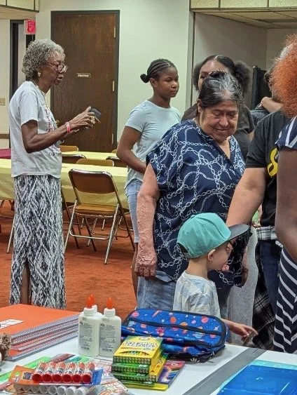 A group of women and a young boy standing in line in a community room, with tables of supplies such as hand sanitizer, notebooks, and scissors in the foreground.