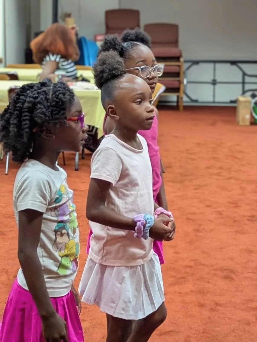 Three young girls standing in a row indoors, attentively watching something. They have different hairstyles and are dressed casually, with two wearing skirts and one wearing a t-shirt.