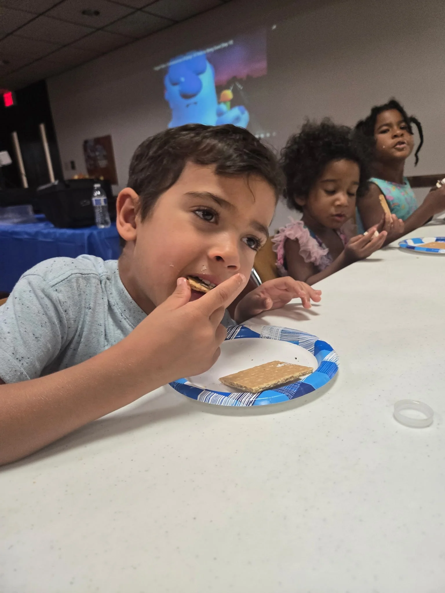 Children sitting at a table eating cookies during a party with a colorful animated movie playing on a screen in the background.