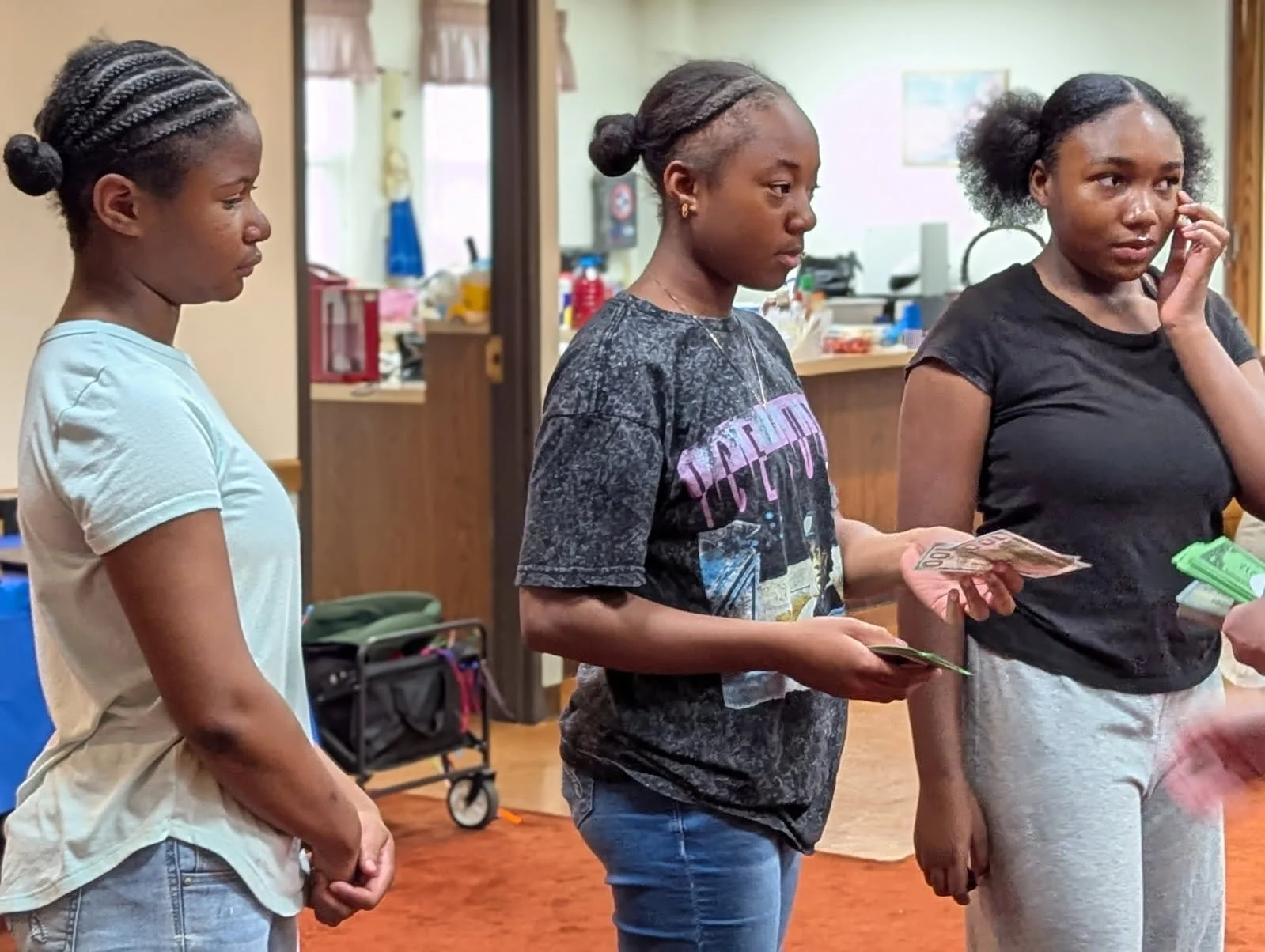 Three young women standing indoors, one holding cash and a card, appearing to be in line or waiting.