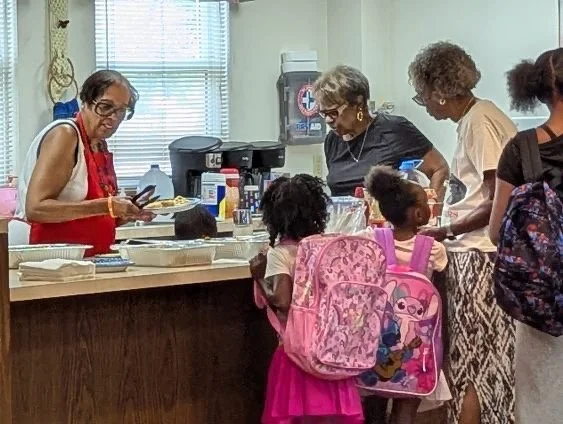 Group of women and children gather at a food serving counter in a community center, with some children carrying pink backpacks and a woman wearing a red apron serving food.