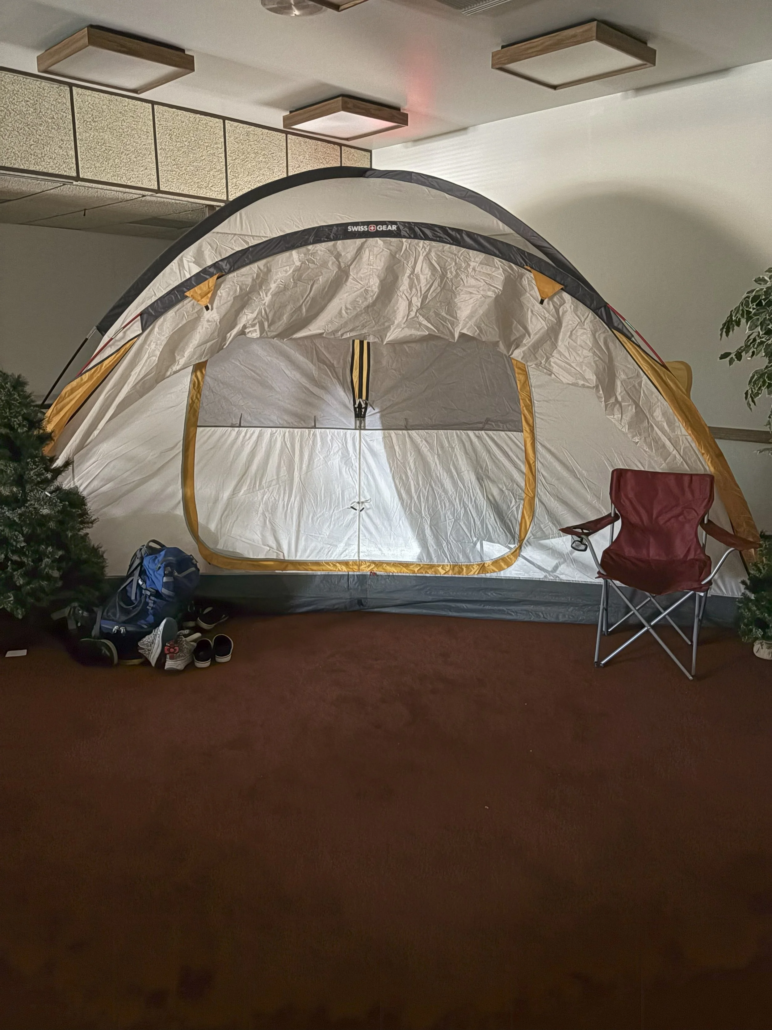 Indoor scene with a large camping tent, a red foldable chair, a blue backpack, and some shoes on a carpeted floor, with ceiling lights above.