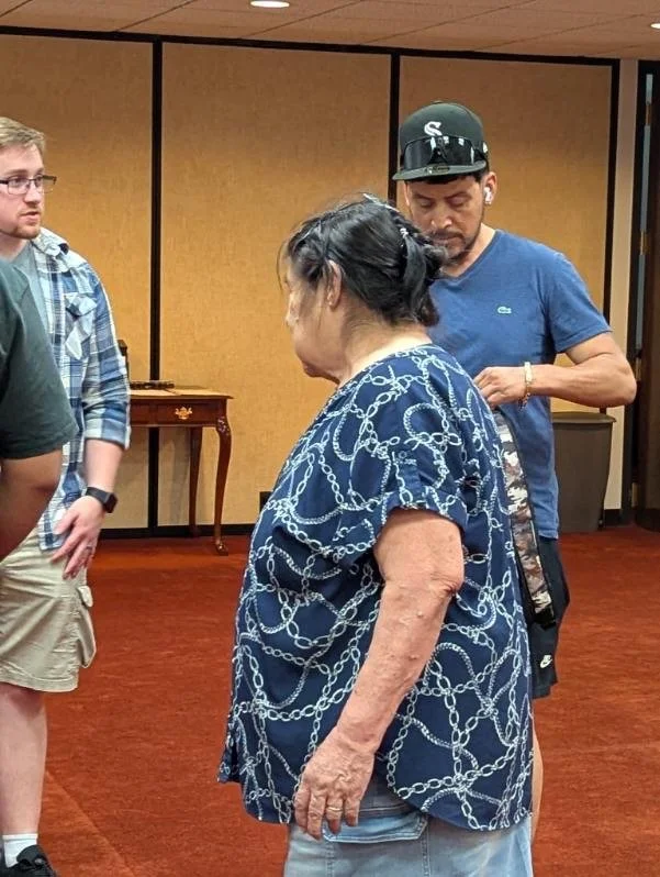 An elderly woman with dark hair pulled back, wearing a blue shirt with a white chain pattern, standing in a room with wood panel walls. A man in a blue t-shirt and cap, and a young man wearing glasses and a plaid shirt are nearby.