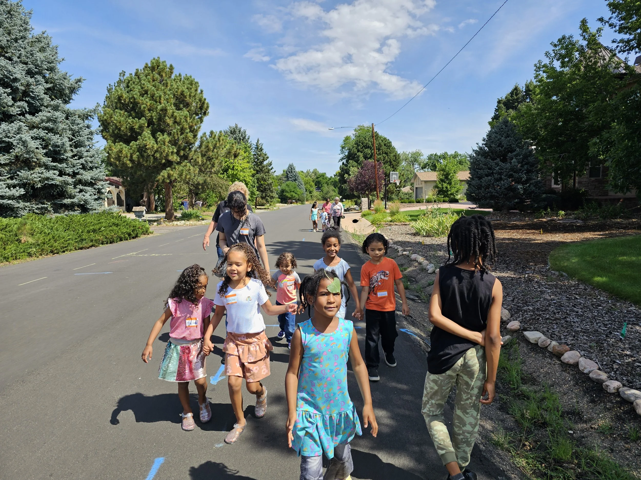 Group of children walking on a suburban street with trees and houses, some with adults, sunny day.