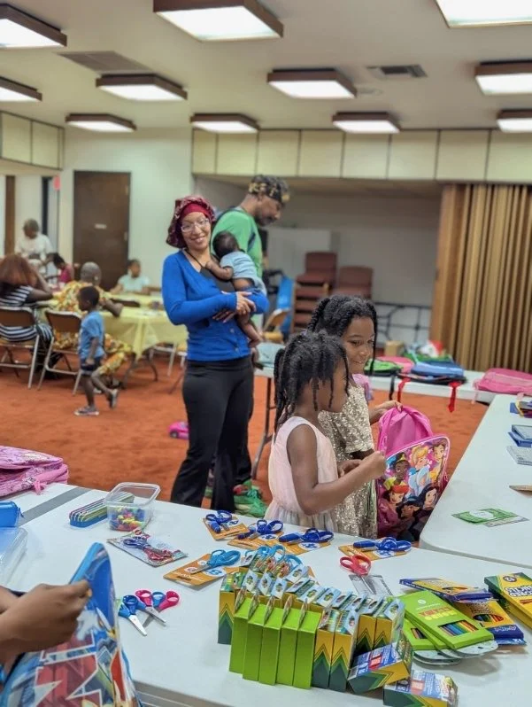 Children and adults at a community event or party, with tables of school supplies and backpacks, in an indoor room with ceiling lights and wood-paneled walls.