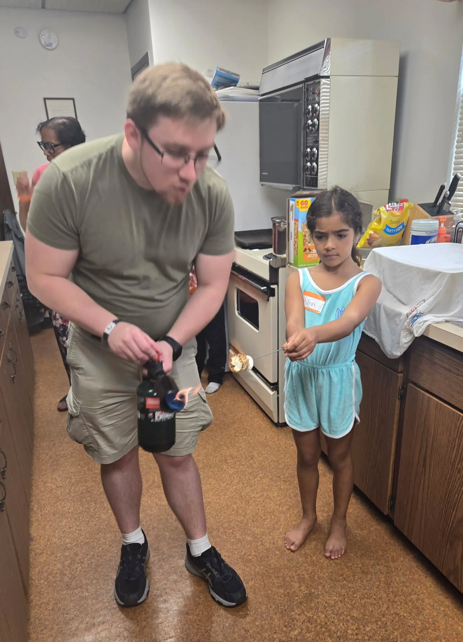 A young girl with a serious expression holding a marshmallow on a stick, while a man in casual clothes is preparing to roast a marshmallow over a flameless butane torch in a kitchen.
