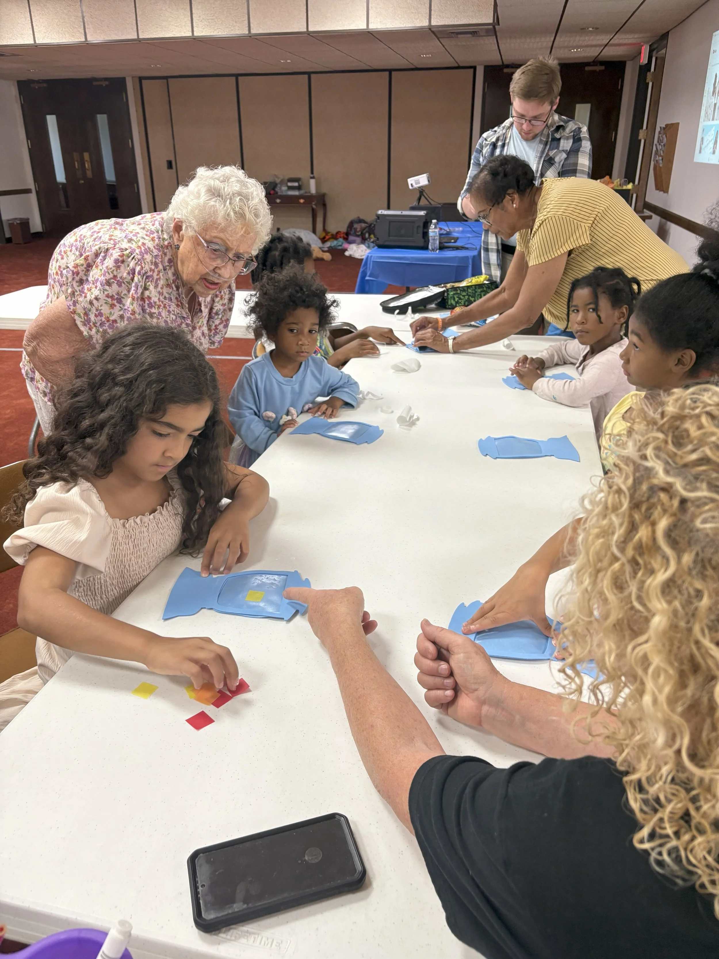 Children and adults gathered around a white table engaging in an arts and crafts activity with colored paper