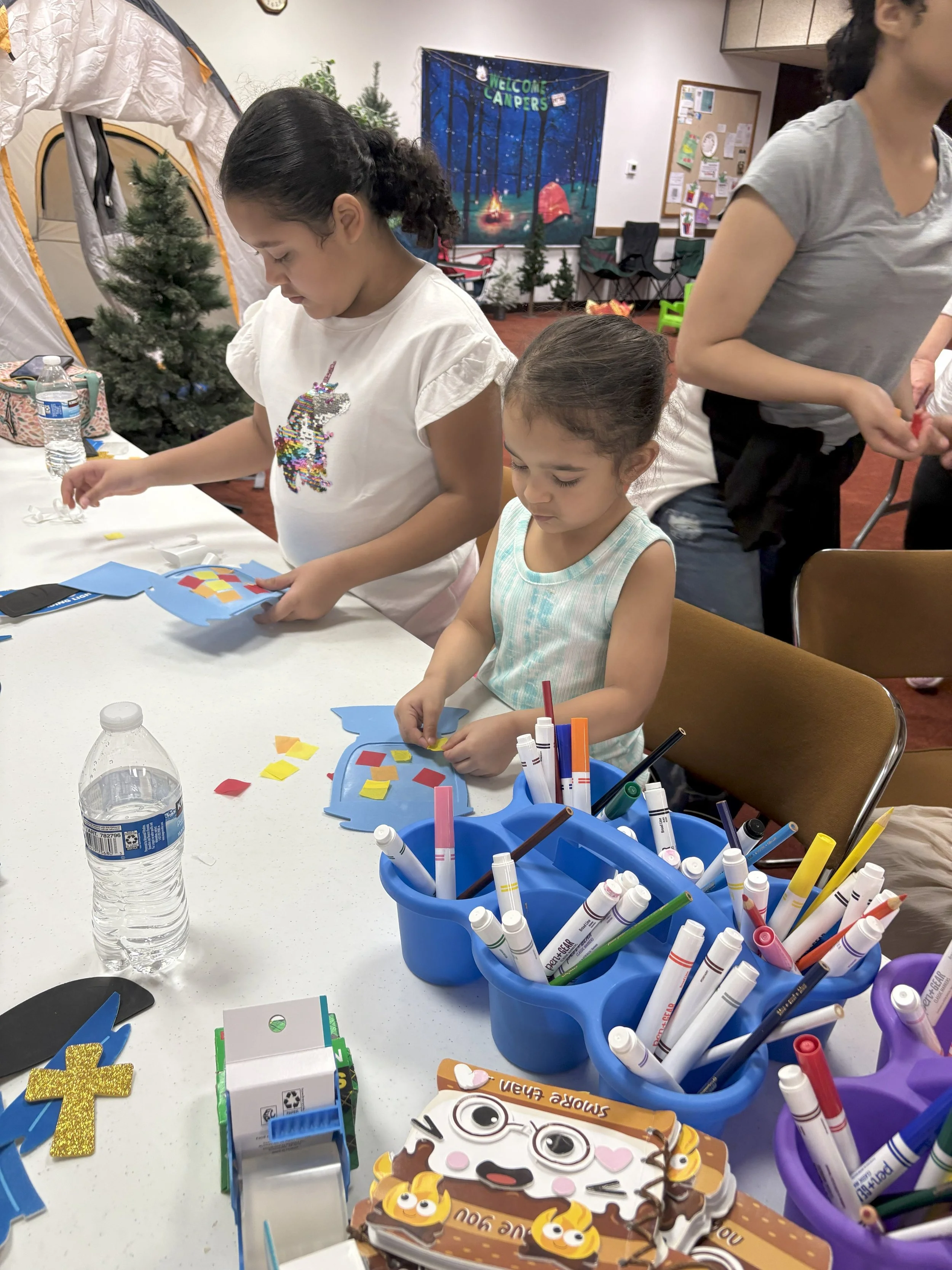 Two young girls decorate paper with stickers at a craft table. One girl with dark hair and a unicorn t-shirt is pasting stickers, while the other girl with brown hair in a blue dress is working with small paper pieces.