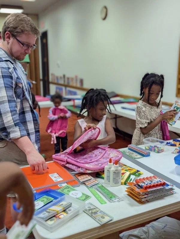 Adults and children at a table with school supplies, including glue, rulers, notebooks, and cash, in a classroom setting.