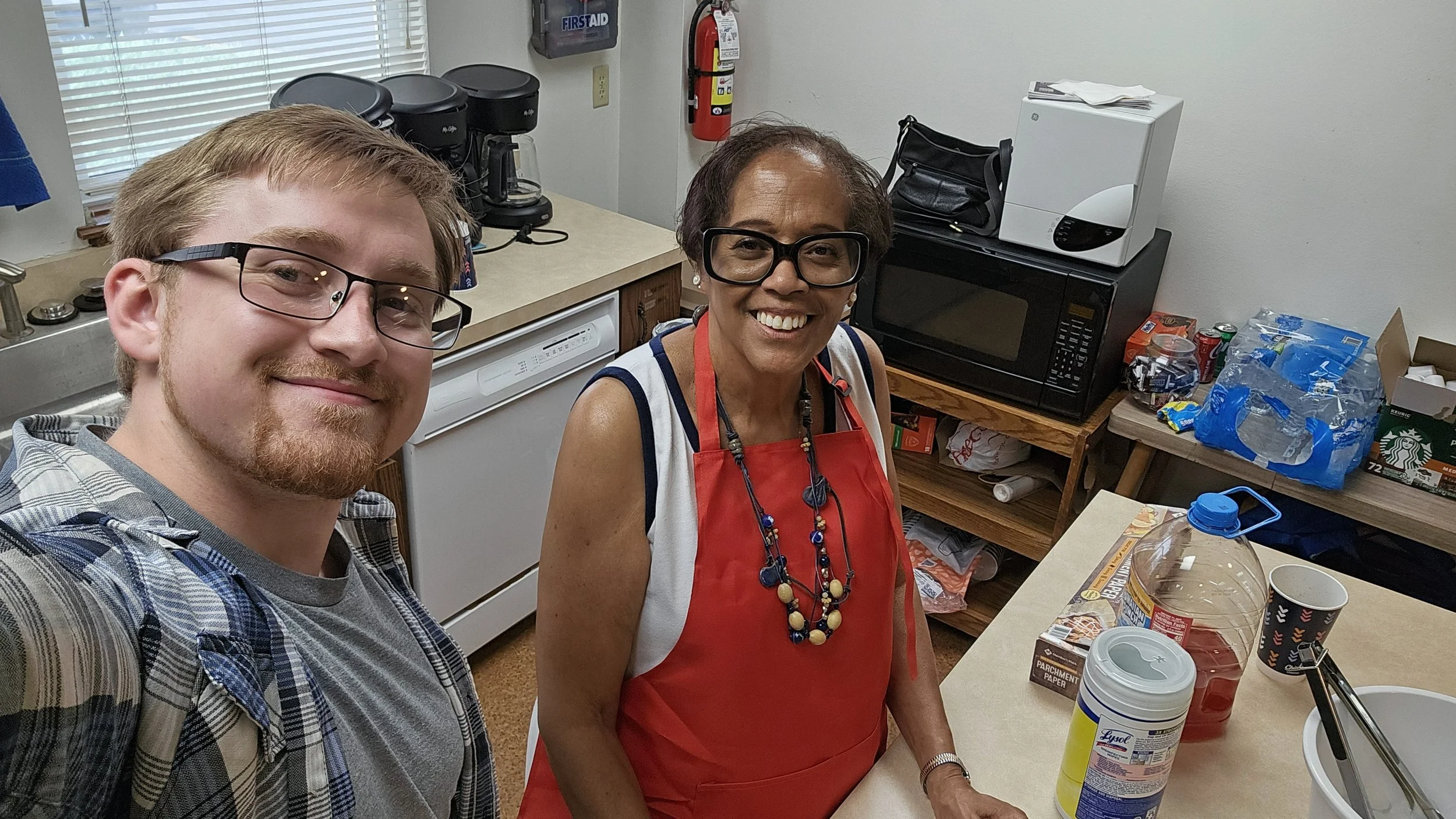 A man and a woman smiling in a kitchen. The woman is wearing a red apron and glasses, and the man is wearing glasses and a plaid shirt. The kitchen has a microwave, coffee maker, and supplies on the counter.
