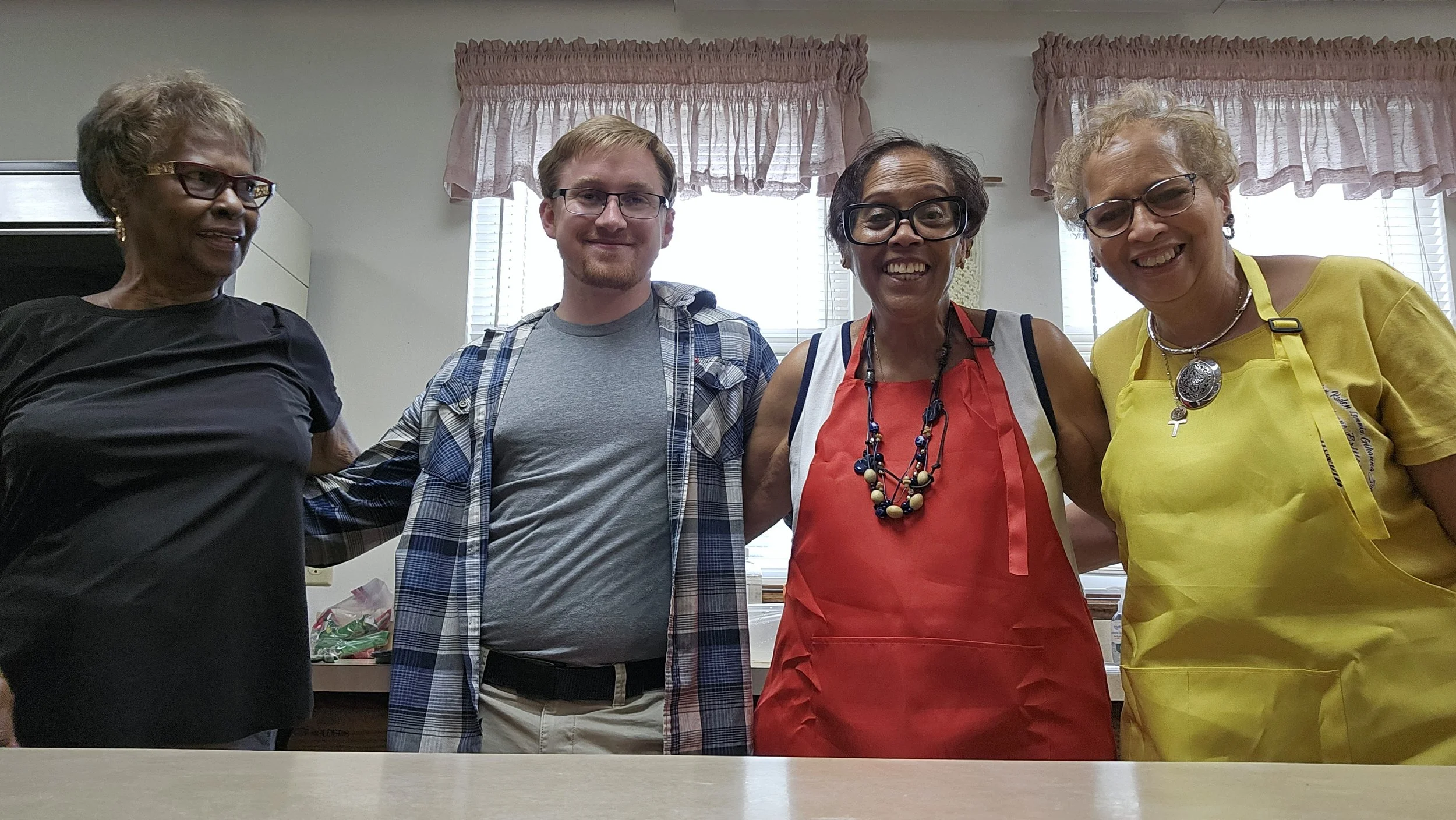Four women and one man standing together inside a room, smiling at the camera. The women are dressed in colorful tops, with two wearing aprons, indicating a possibly celebratory or social event.