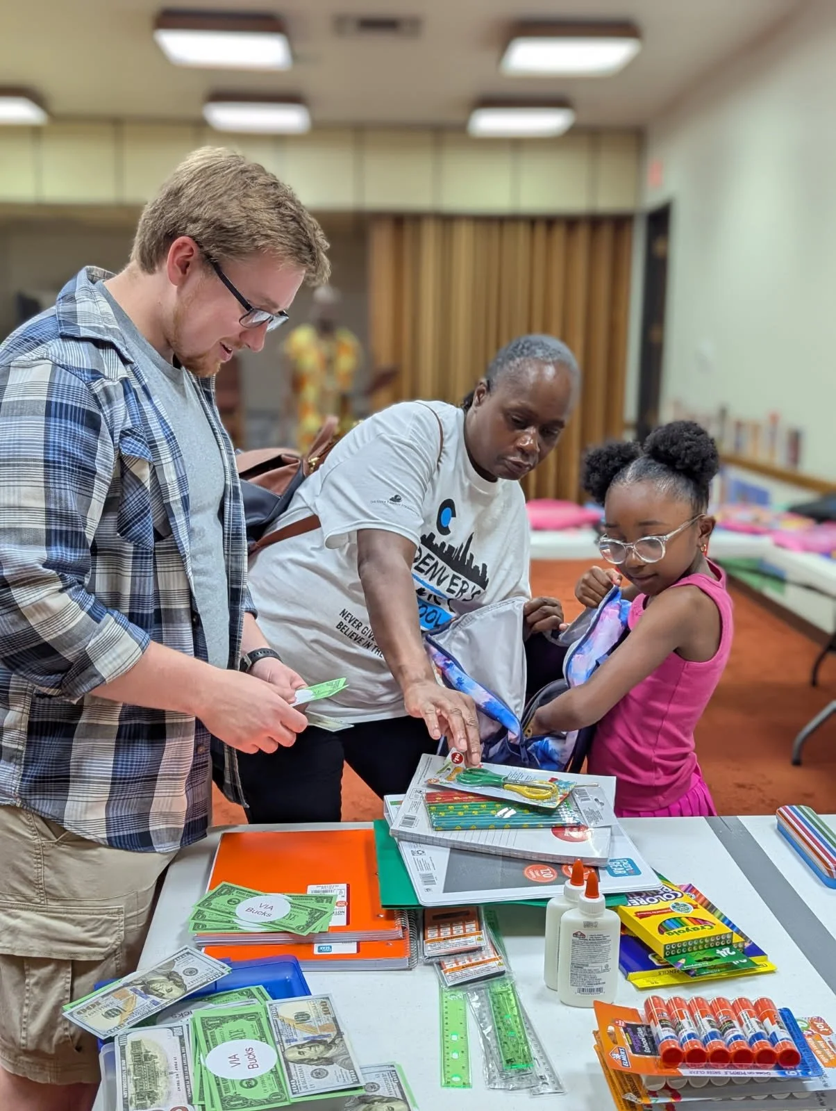 Three people, two adults and a young girl, standing at a table filled with school supplies and money, in a room with fluorescent lighting and a bookshelf in the background.
