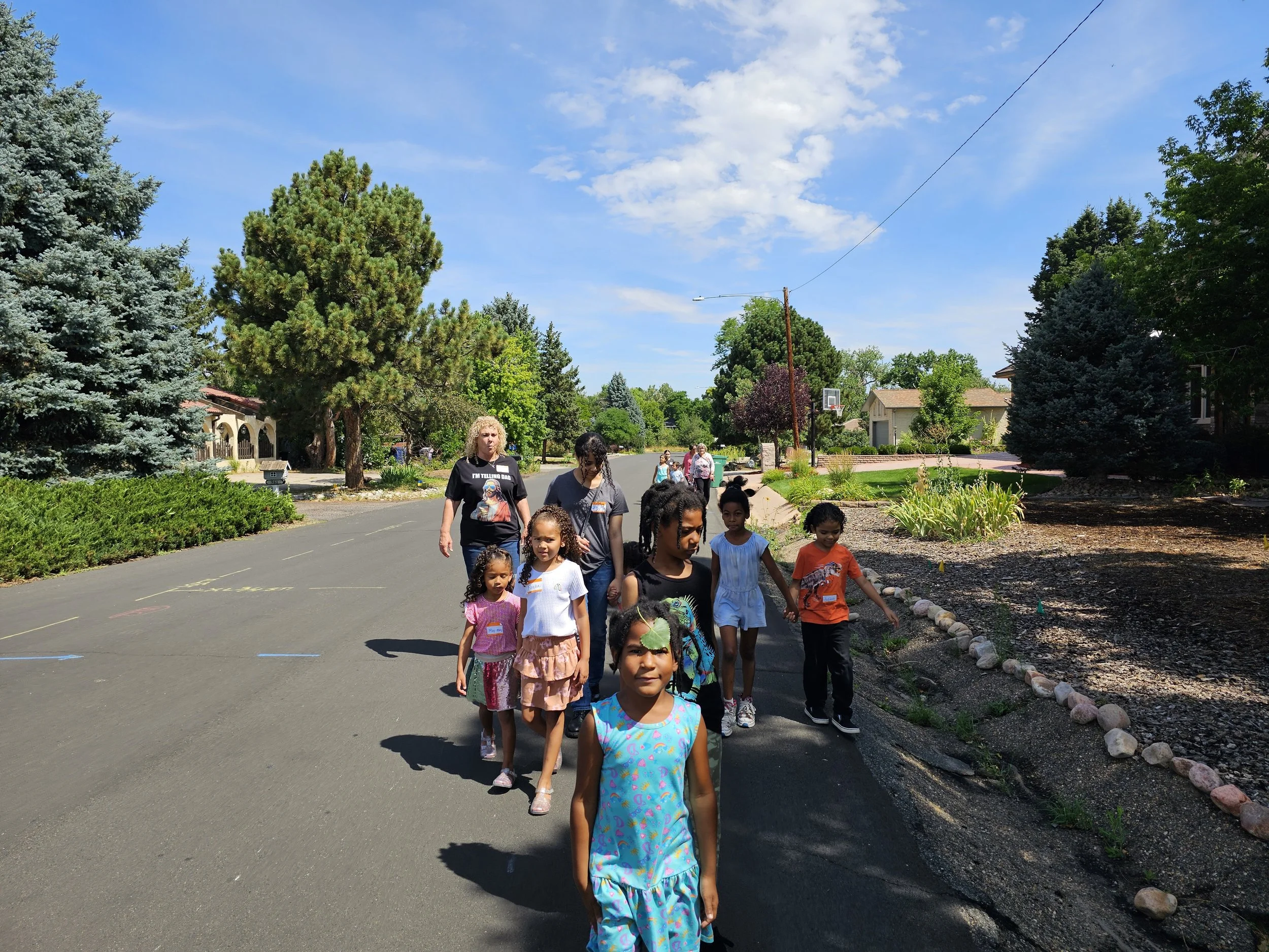 A group of children and adults walking on the sidewalk in a suburban neighborhood on a sunny day.