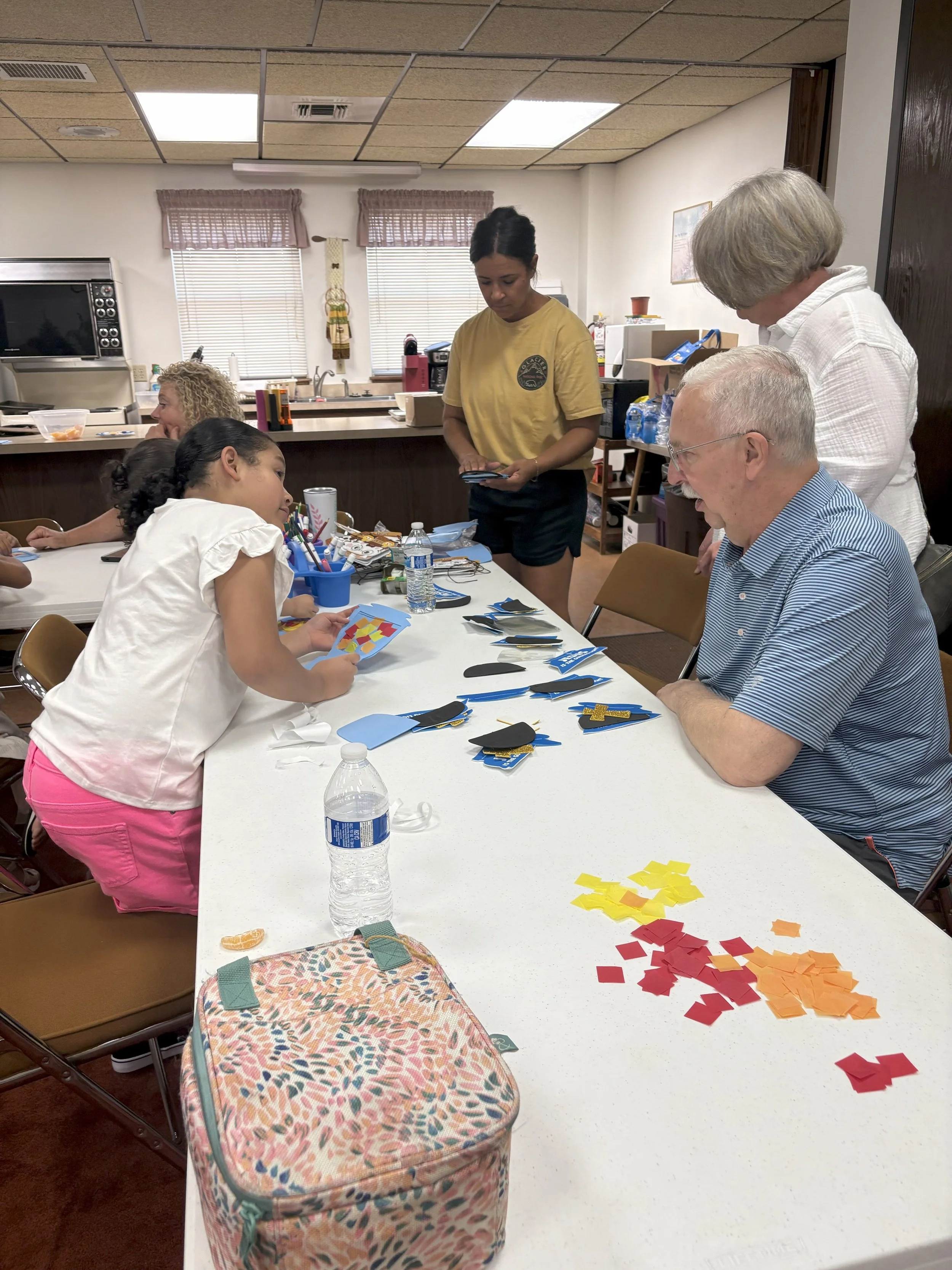 Children and adults sitting around a table making crafts with colored paper, scissors, and other supplies in a kitchen/dining room.