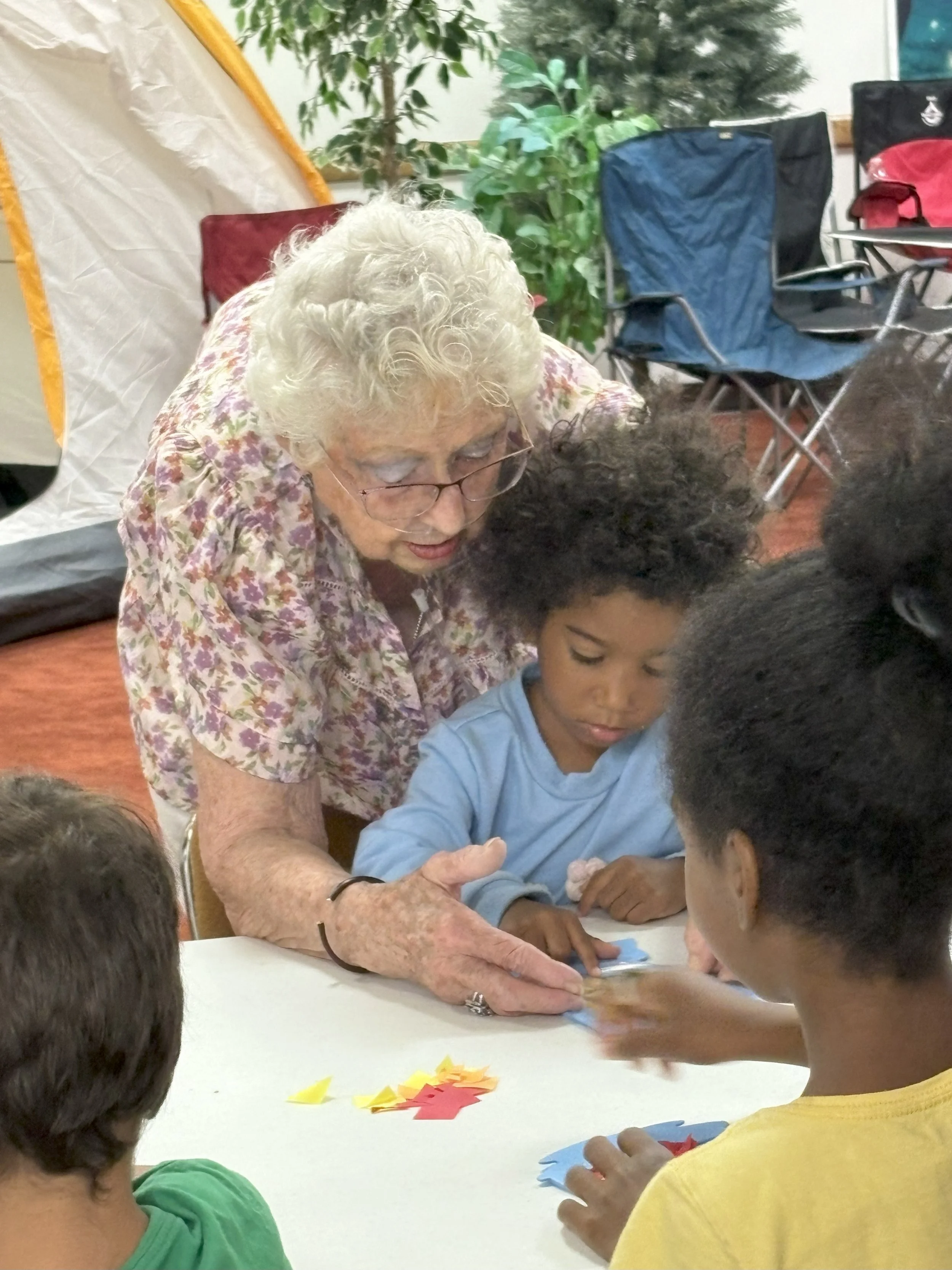 An elderly woman with white hair and glasses helping children with arts and crafts at a table in a community room.