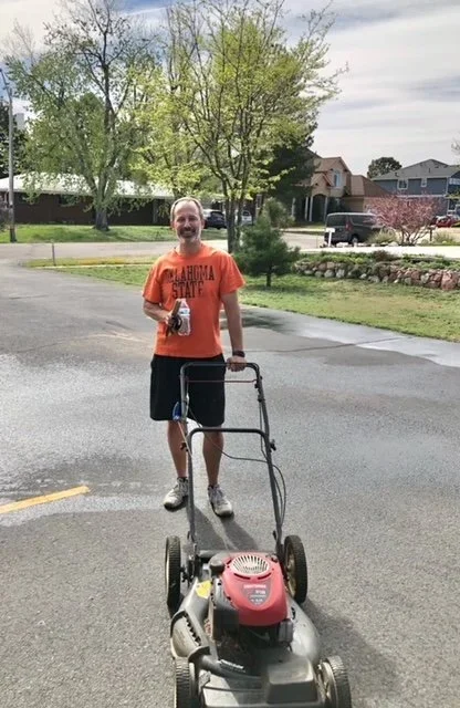 A smiling man with a beard wearing an orange Oklahoma State shirt and black shorts using a grass trimmer in a residential neighborhood with trees and houses.