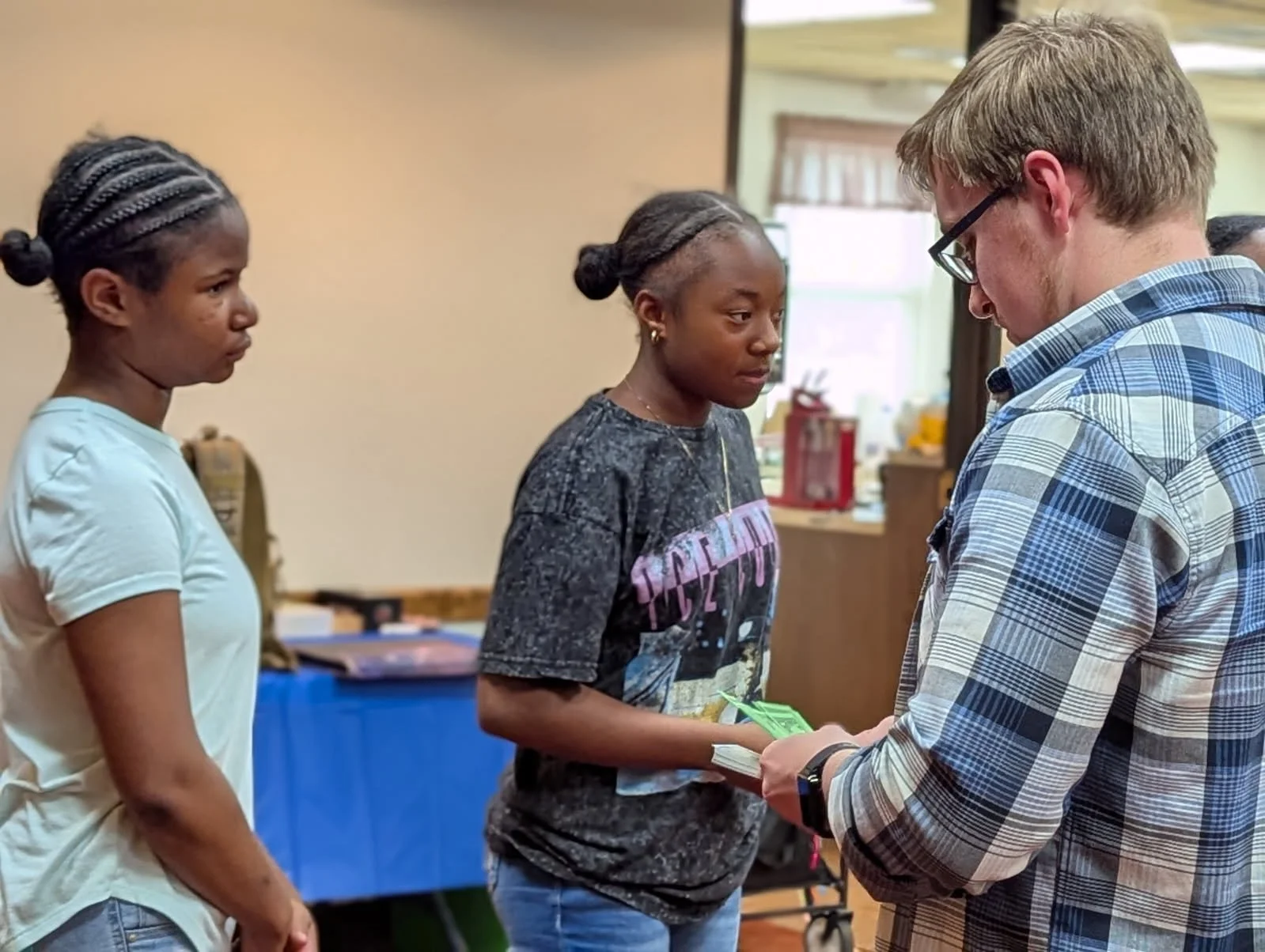Two young women standing in line, receiving money from a man in a plaid shirt, inside a room with a table and window, daytime.
