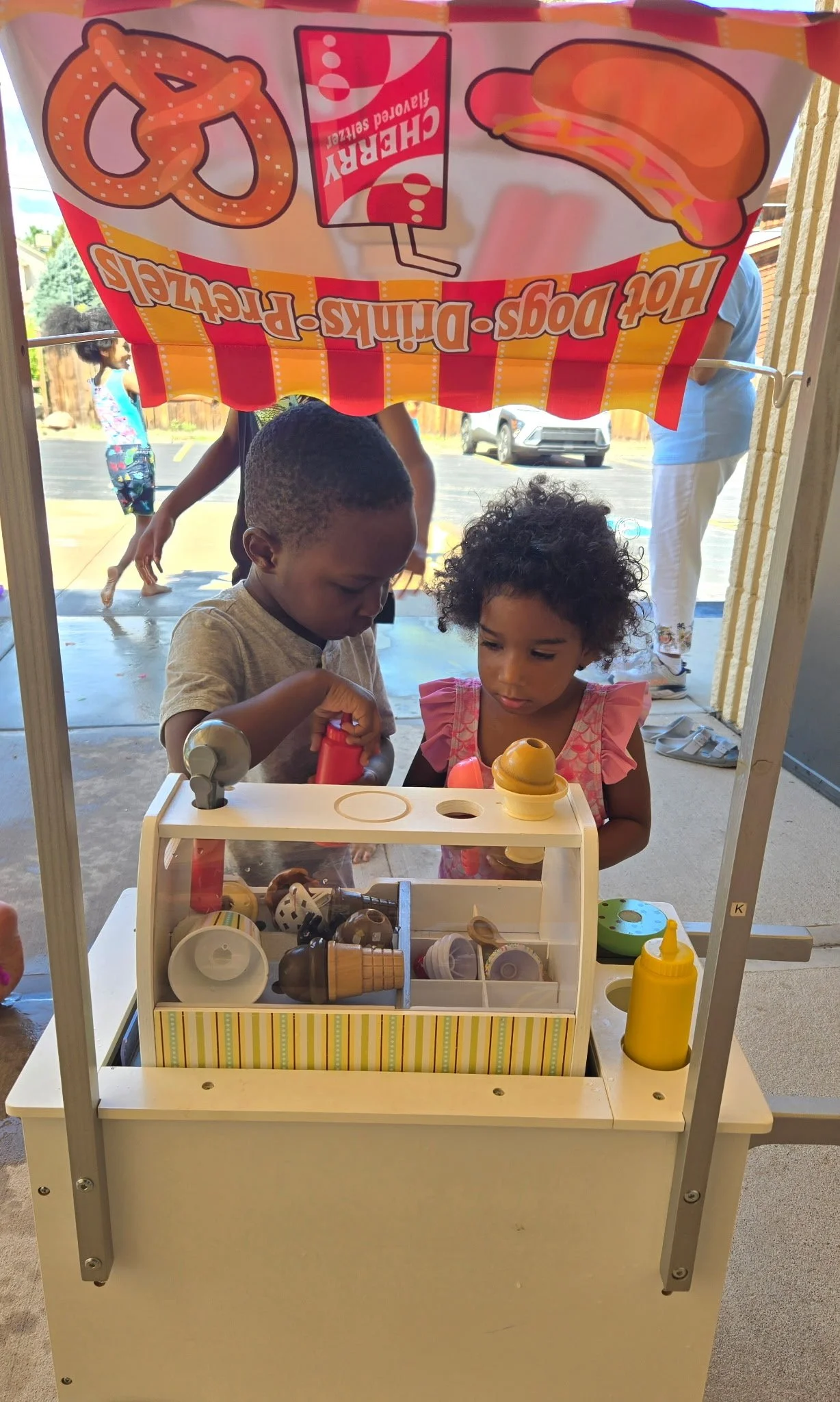 Two children playing with a toy ice cream and hot dog stand, with a colorful banner above advertising pretzels, hot dogs, and fries, outside on a sunny day.