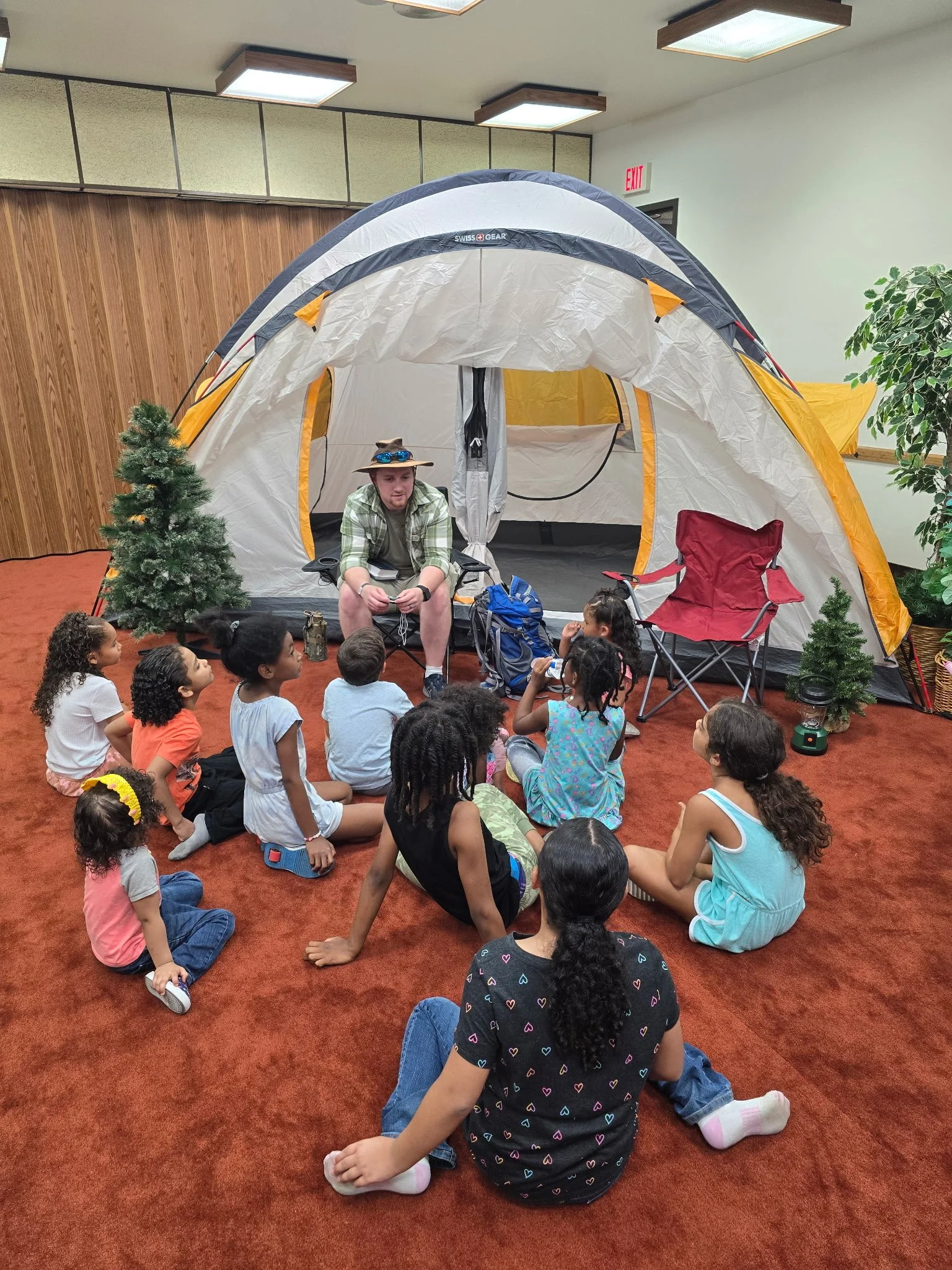 A man is speaking to a group of children seated on the carpet, in front of a camping tent indoors. The children are listening attentively, and the scene is decorated with small artificial trees and camping gear.