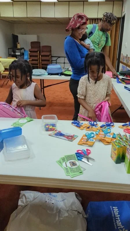 Two young girls are at a table with school supplies, while an adult woman and man stand behind them, looking on in a room with chairs and tables.