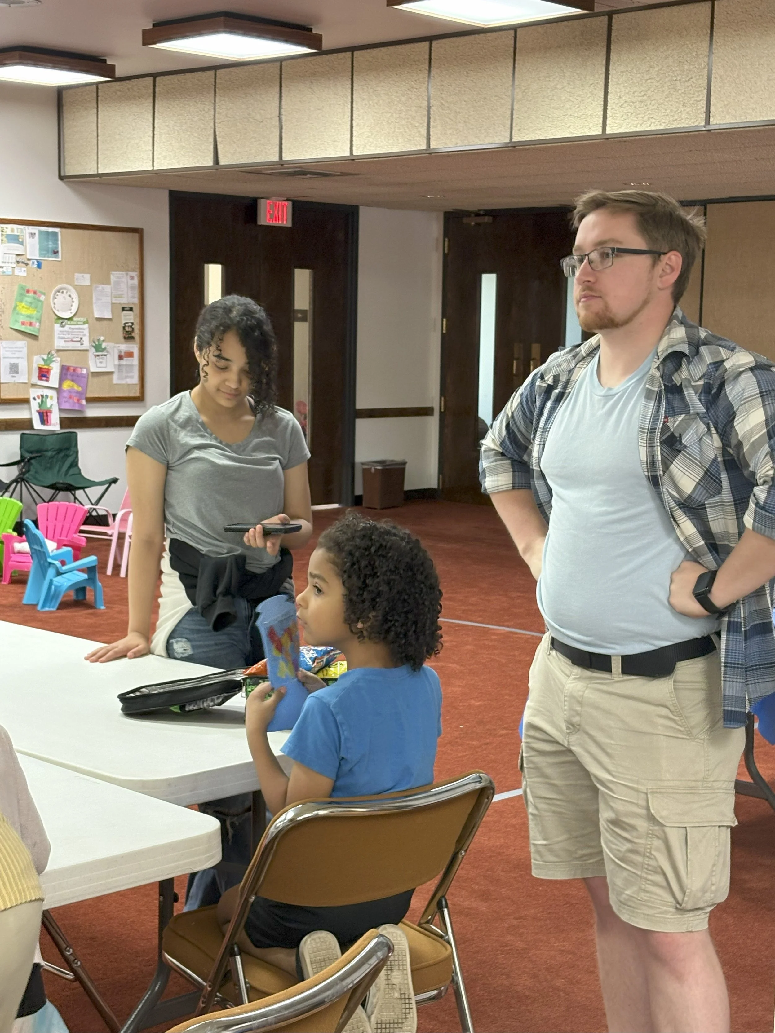 A group of three people, two adults and one child, in a room with red carpet and colorful children's furniture. The woman with curly hair is holding a phone, the young girl is sitting at a table holding a card, and the man with glasses and a plaid sh