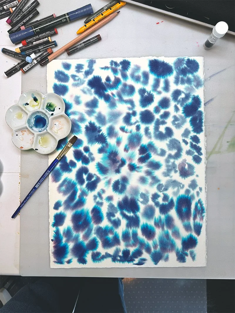 Looking over the top of an artist desk. A piece of paper that has been painted with blue and turquoise watercolor ink.
