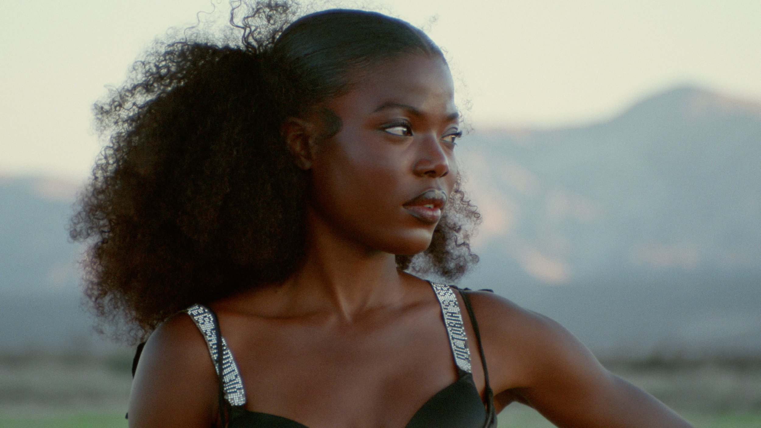 A young African American woman with curly hair styled in a half-up, half-down look, wearing a black tank top with silver straps, gazing thoughtfully into the distance outdoors during a sunset.