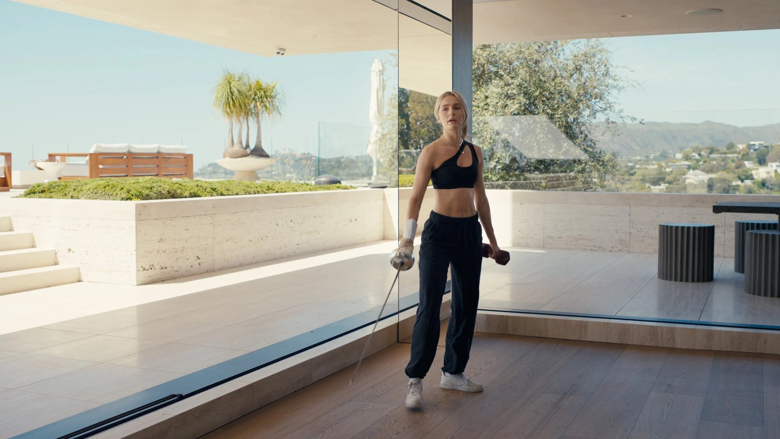 A woman in workout clothes holding dumbbells in one hand and a resistance band in the other, standing inside a modern house with large glass windows and a scenic mountain view outside, film emulation color grade