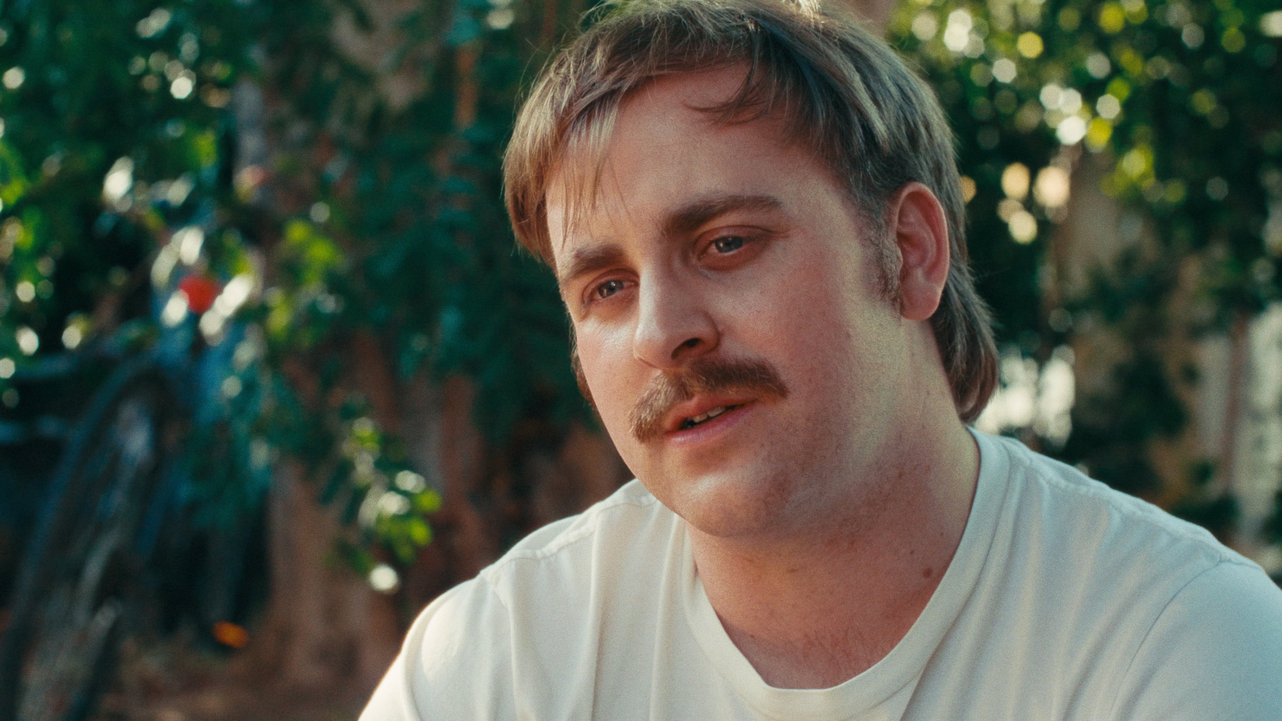 A man with light brown hair, a mustache, and light skin wearing a white shirt outdoors with green foliage in the background, vintage film preset colors