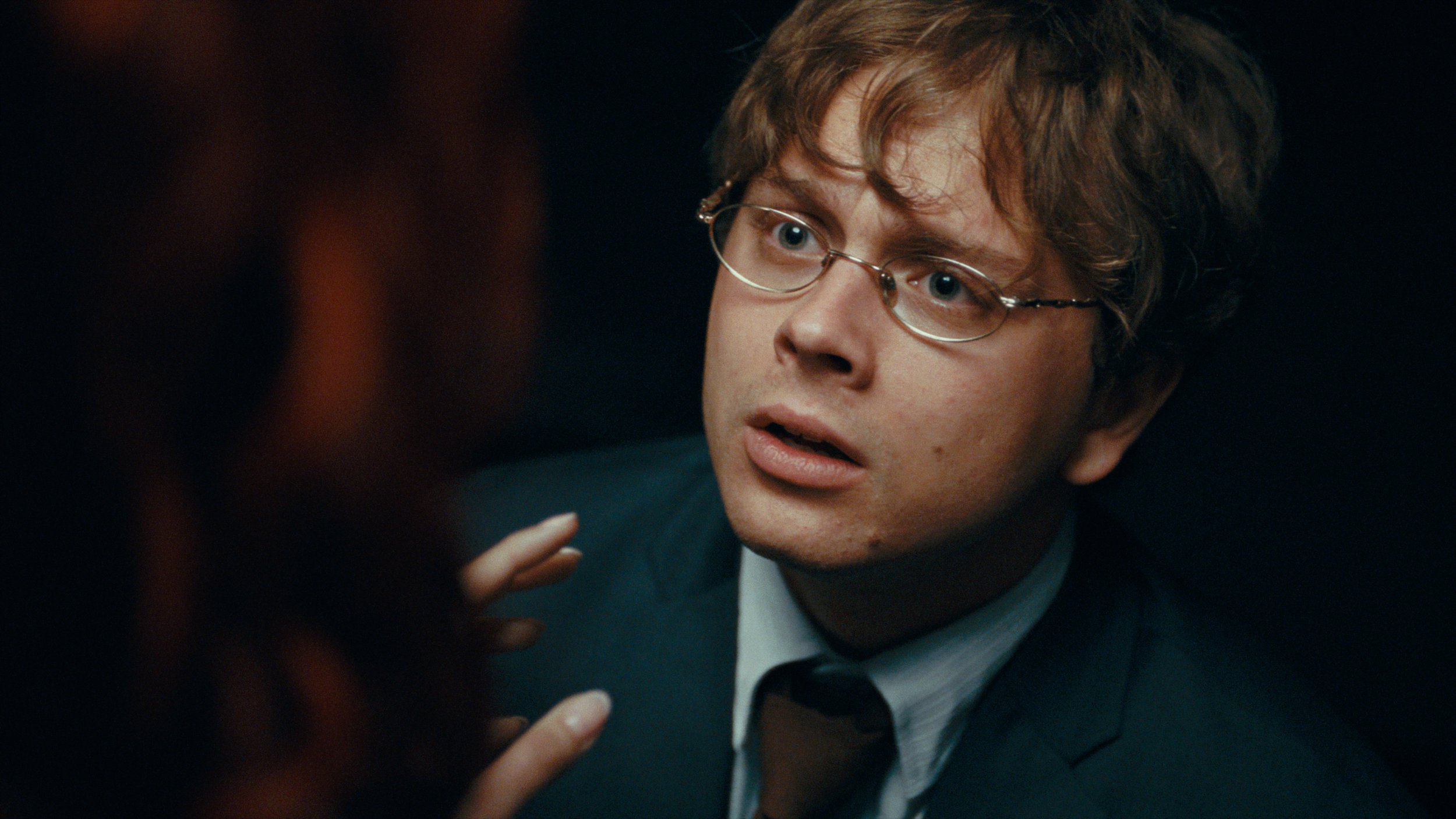 A young man with glasses and curly red hair, wearing a dark jacket and white shirt, appears surprised or intrigued in a dark setting.