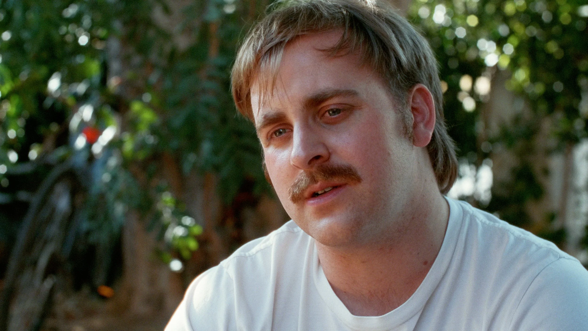 A young man with light brown hair and a mustache looking down and to the side, outdoors with greenery in the background, 35mm film look with grain