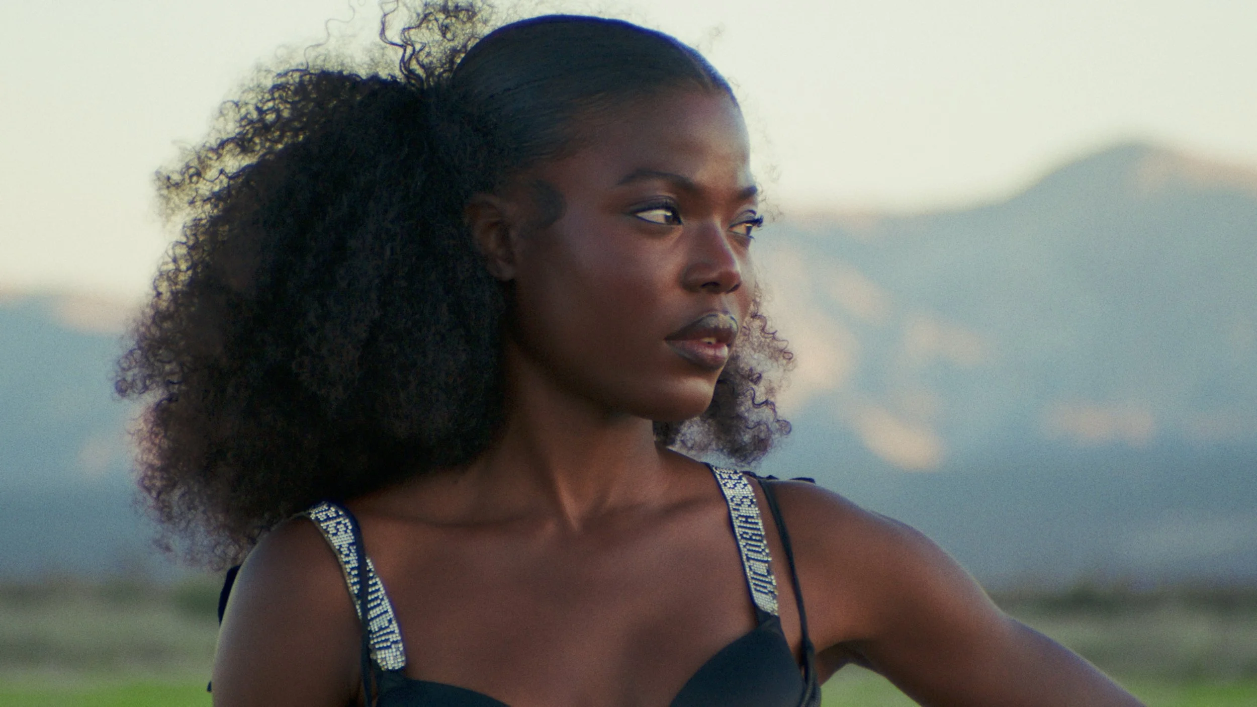 A woman with dark skin and curly hair looks to the side against a background of sky and clouds.