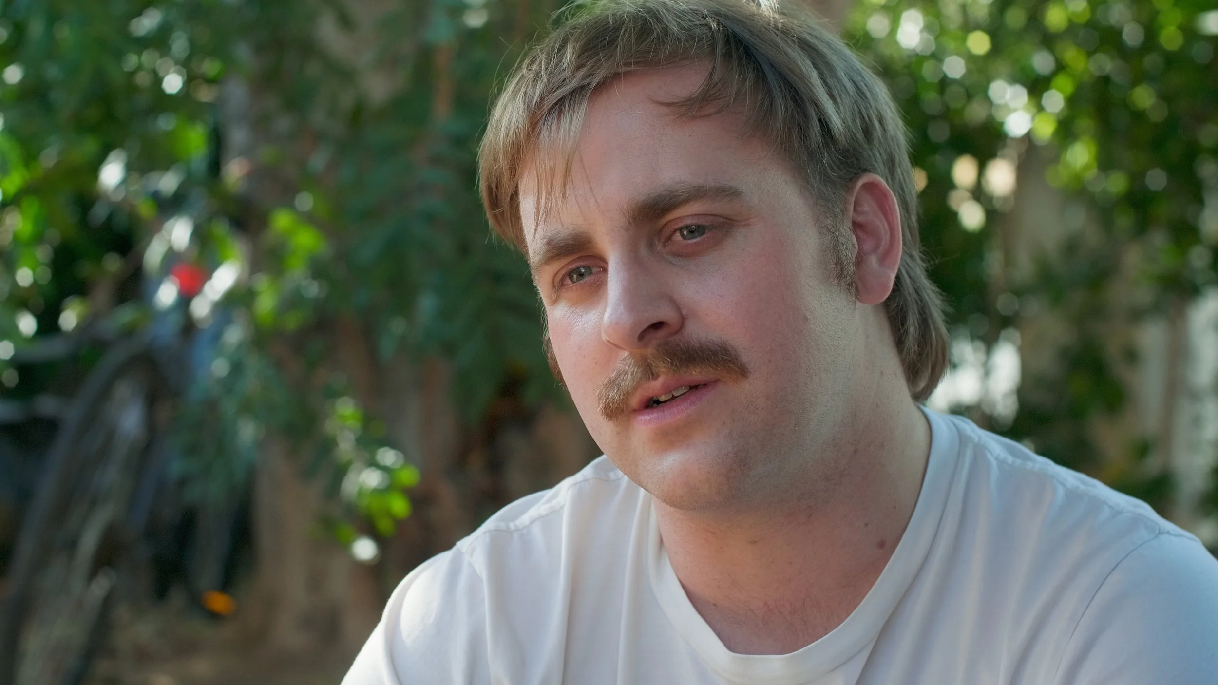 A young man with light brown hair, a mustache, and a beard wearing a white T-shirt outdoors with green foliage in the background