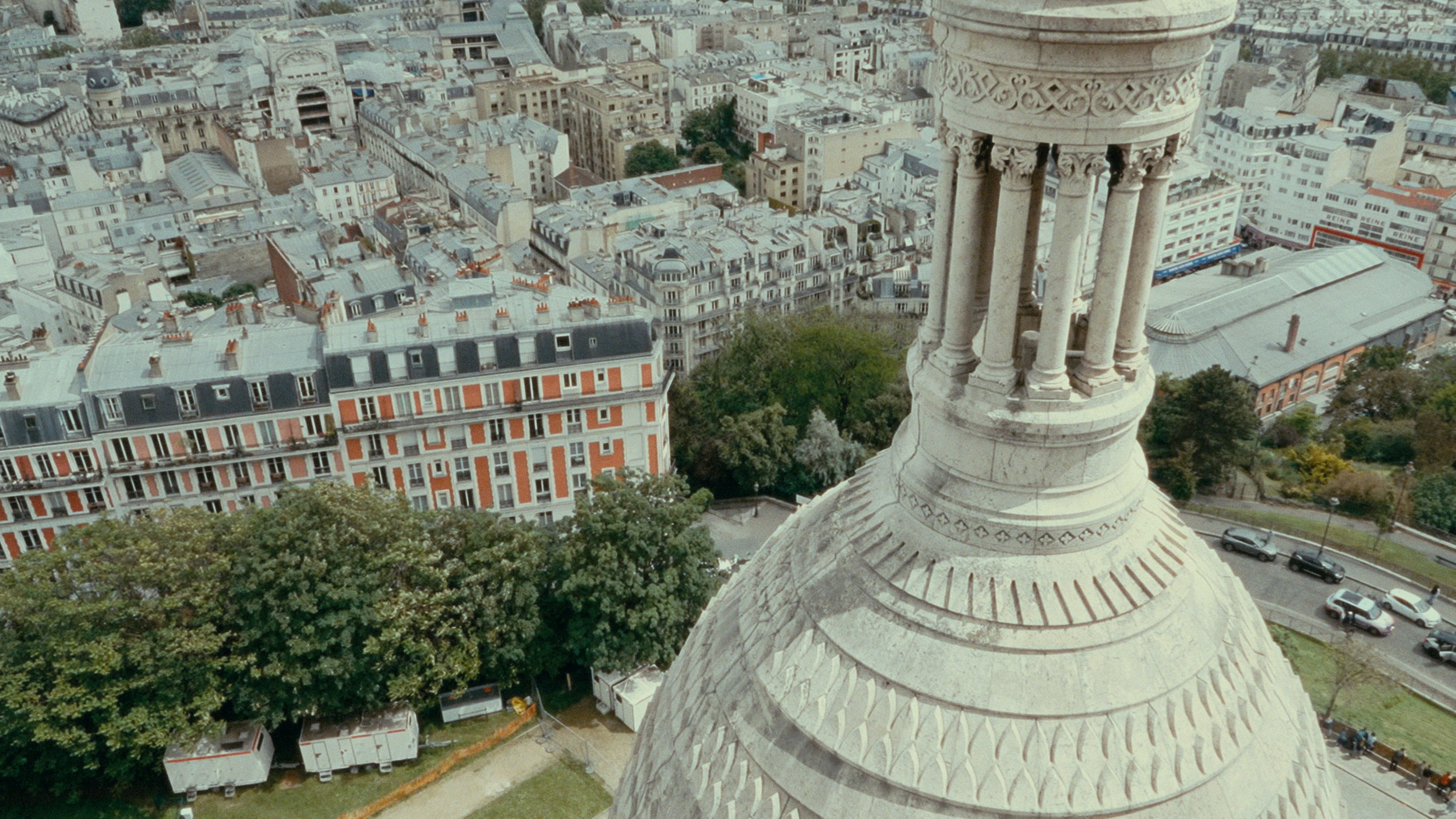 View from a church tower showing the city of Paris with rooftops, buildings, and trees, and a partial view of the church’s ornate dome in the foreground.