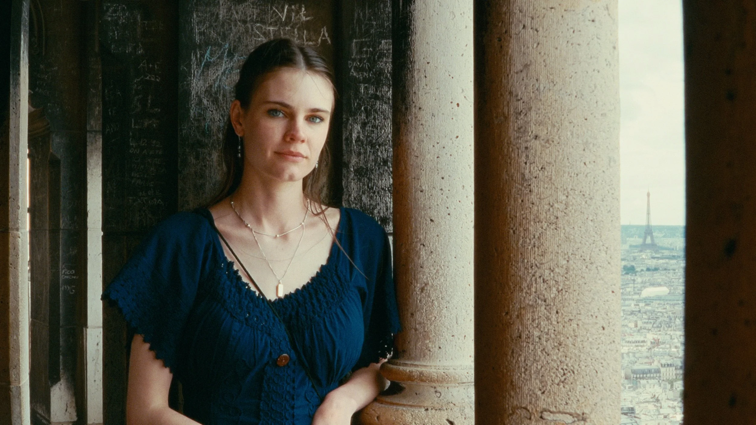 A young woman with brown hair and blue eyes standing on a balcony of a tall building in Paris with the Eiffel Tower visible in the background, kodak film emulation color grade