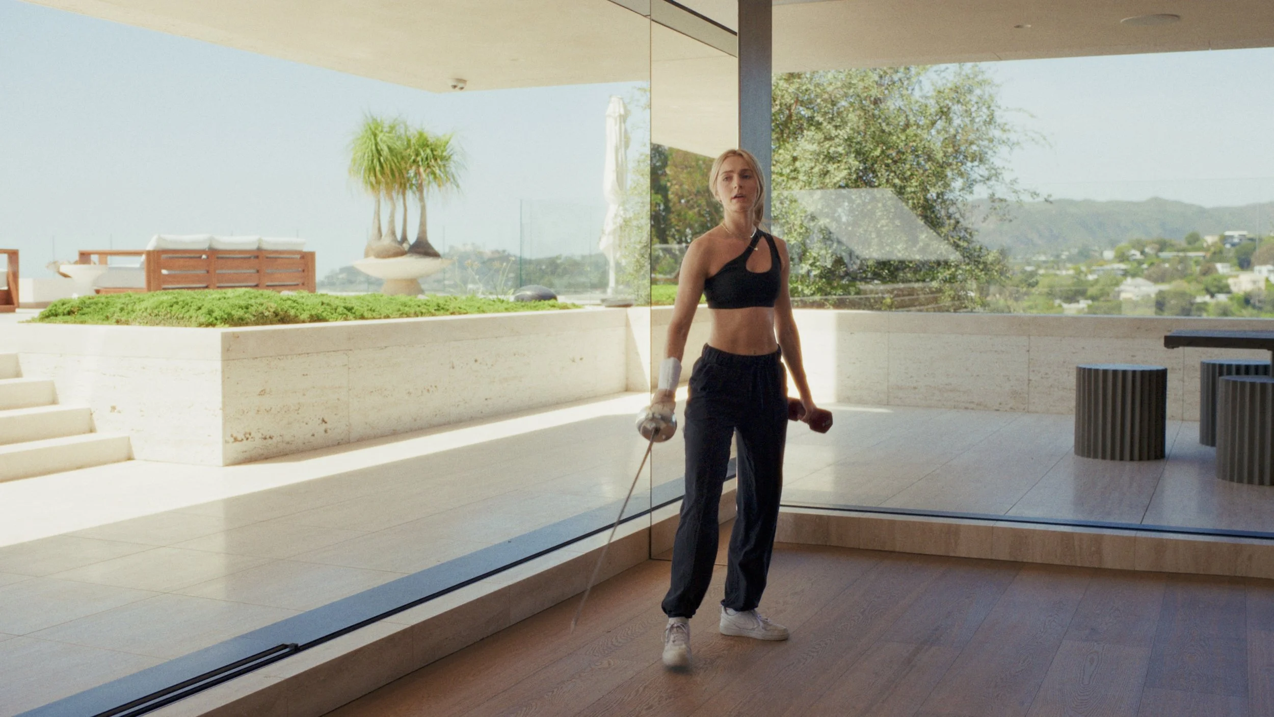 A young woman in athletic clothing walking with a white dumbbell in her right hand, inside a modern building with large glass windows overlooking a scenic outdoor view.