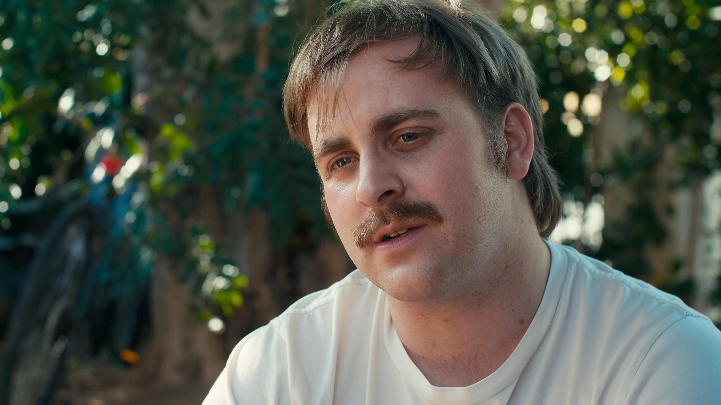 A close-up of a man with light brown hair and a mustache, wearing a white shirt, outdoors with blurred green foliage in the background.