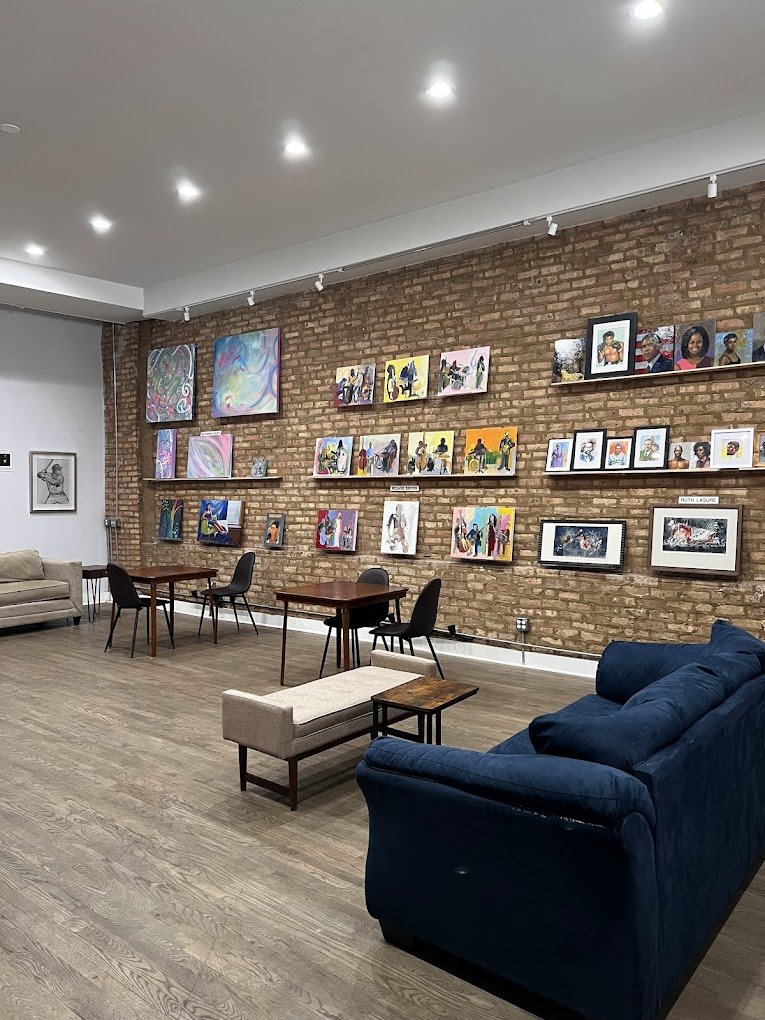An art gallery with a brick wall displaying colorful paintings, a blue couch, a beige bench, small tables, and black chairs in a well-lit room.