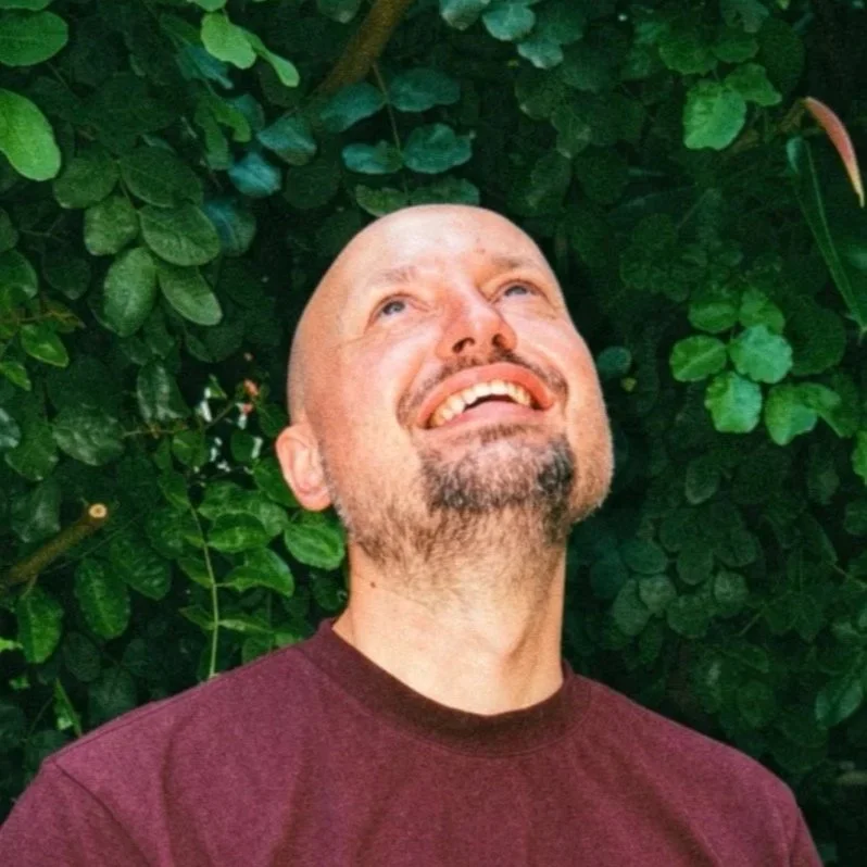 A bald man with a beard looking up and smiling, standing in front of green foliage.
