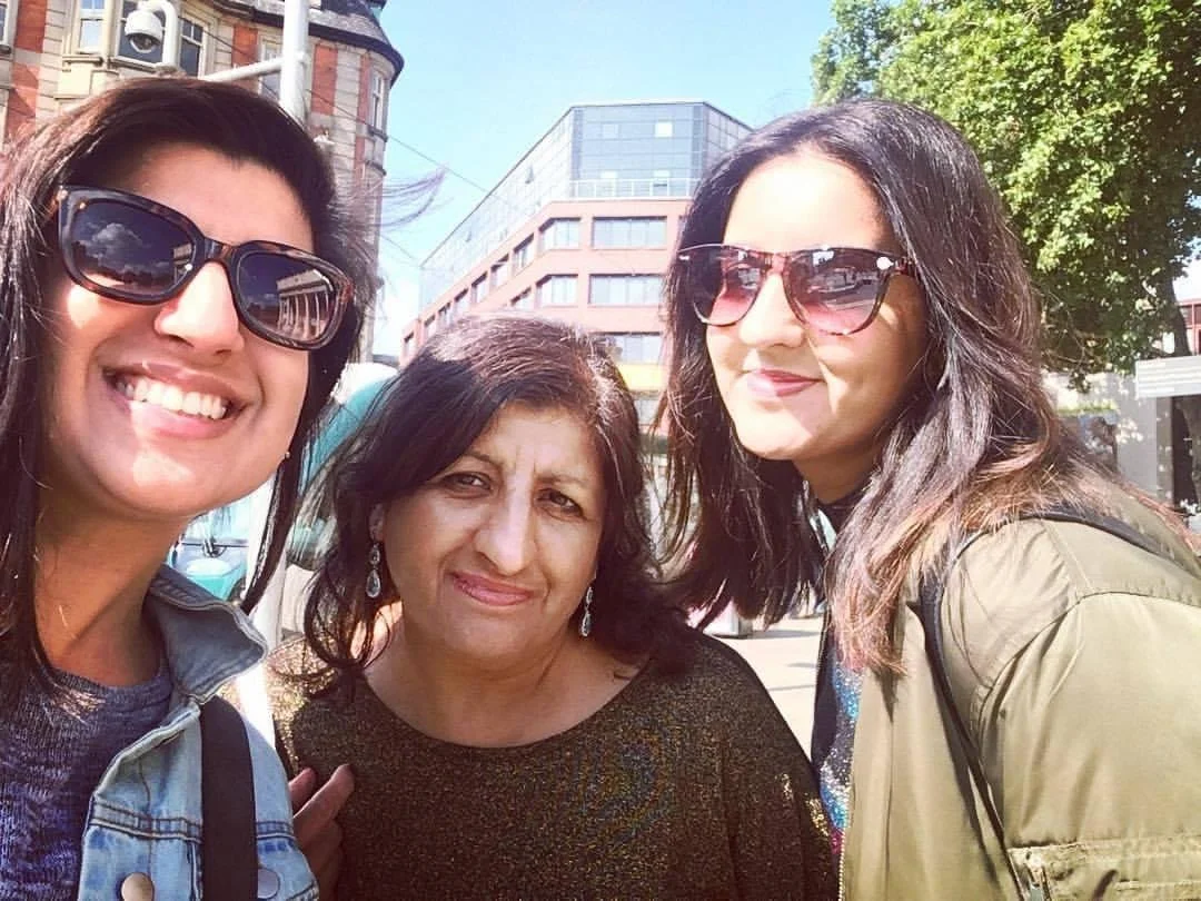 Mandy, her mom and her sister smiling at the camera. They are wearing sunglasses and standing in front of buildings.