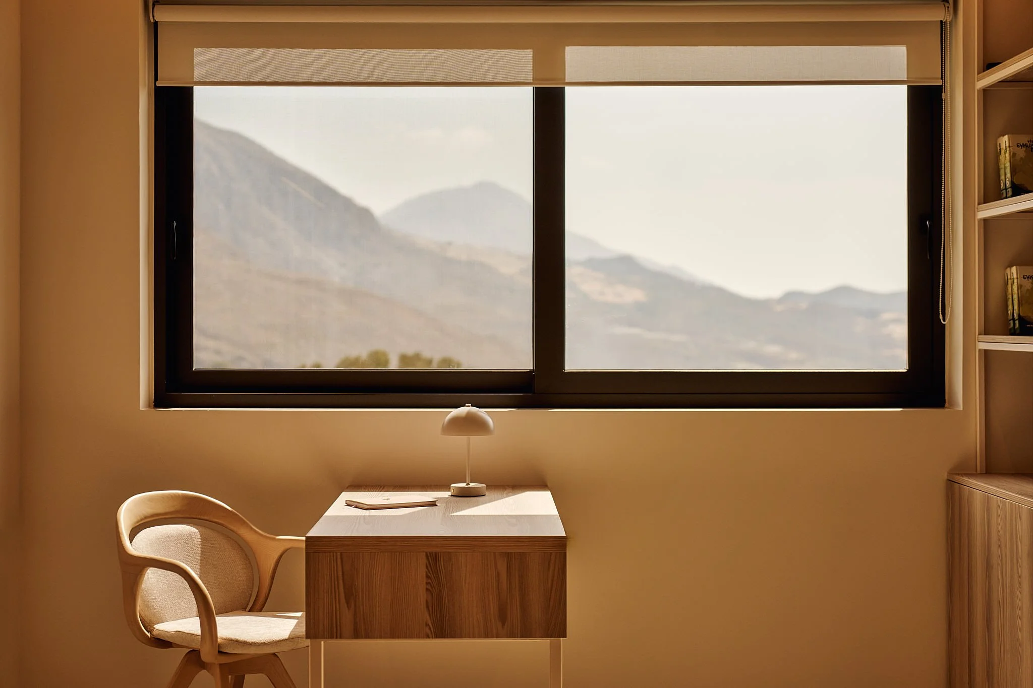 A minimalist workspace with a wooden desk, a beige upholstered chair, a small white desk lamp, and a large window with a view of mountains in the distance.